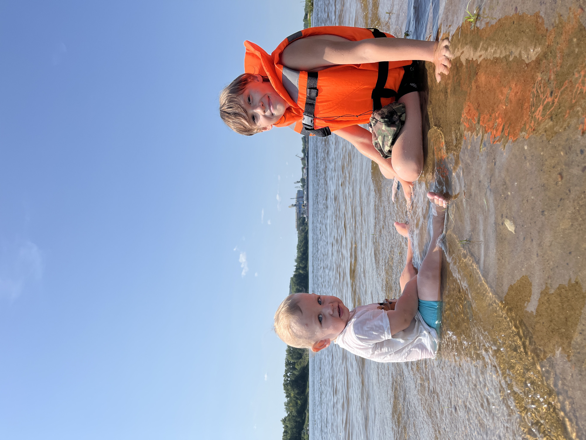 Two children are sitting in shallow water on a sandy shore with a river and trees in the background under a clear blue sky. The child on the right, appearing to be older, is wearing an orange life vest and smiling at the camera. The child on the left, younger, is looking away from the camera. The image was taken on the Volga River embankment in Nizhny Novgorod, Russia.