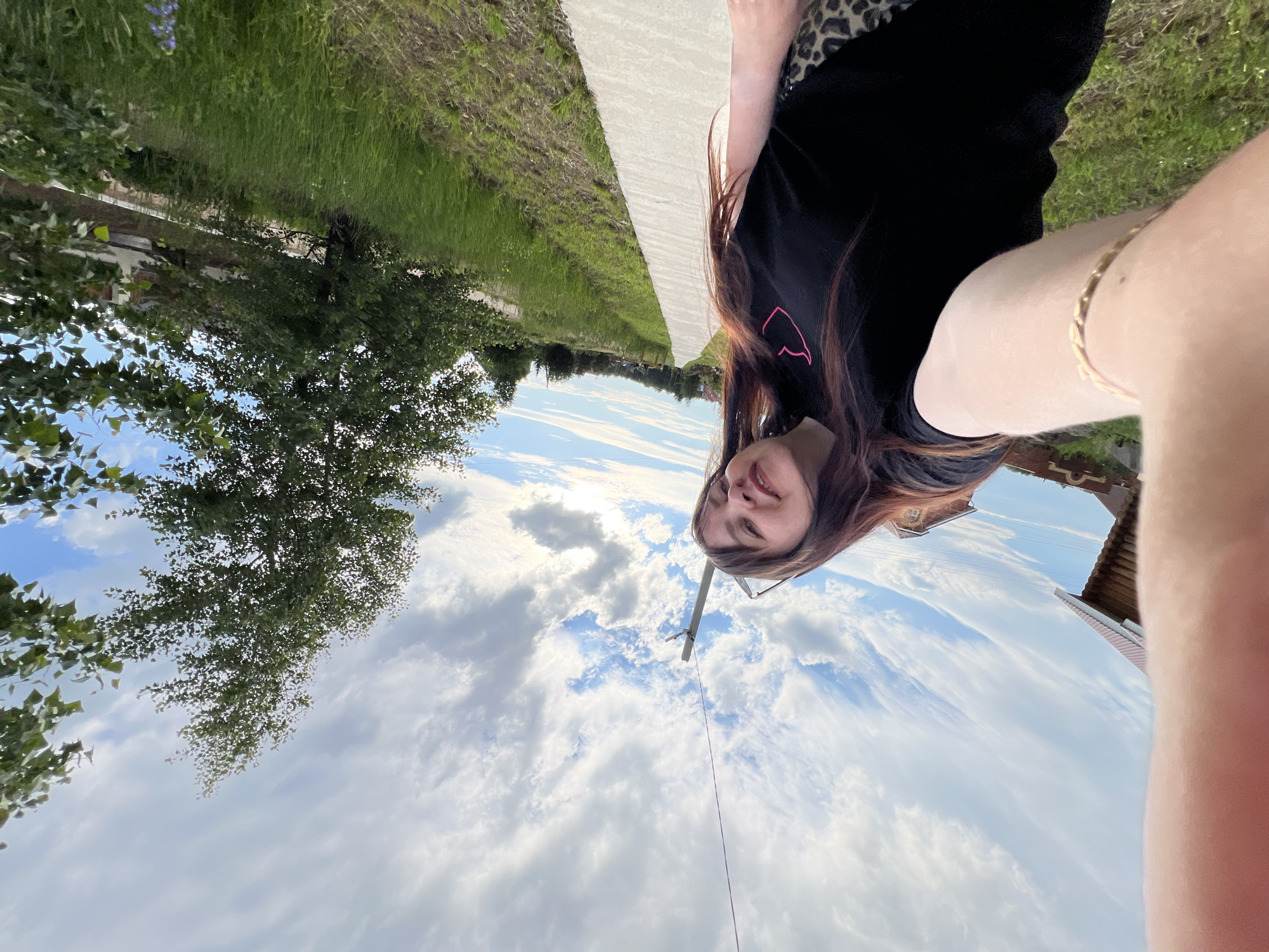 A young woman with long, dark hair is taking a selfie outdoors. She is wearing a black t-shirt with a pink heart graphic and leopard print bottoms. She has a bracelet on her left wrist. The woman is standing on a concrete pathway that runs along a grassy area. A utility pole and wires are visible in the background, along with trees and houses. The sky is partly cloudy. The location is identified as Noviy, Russia.