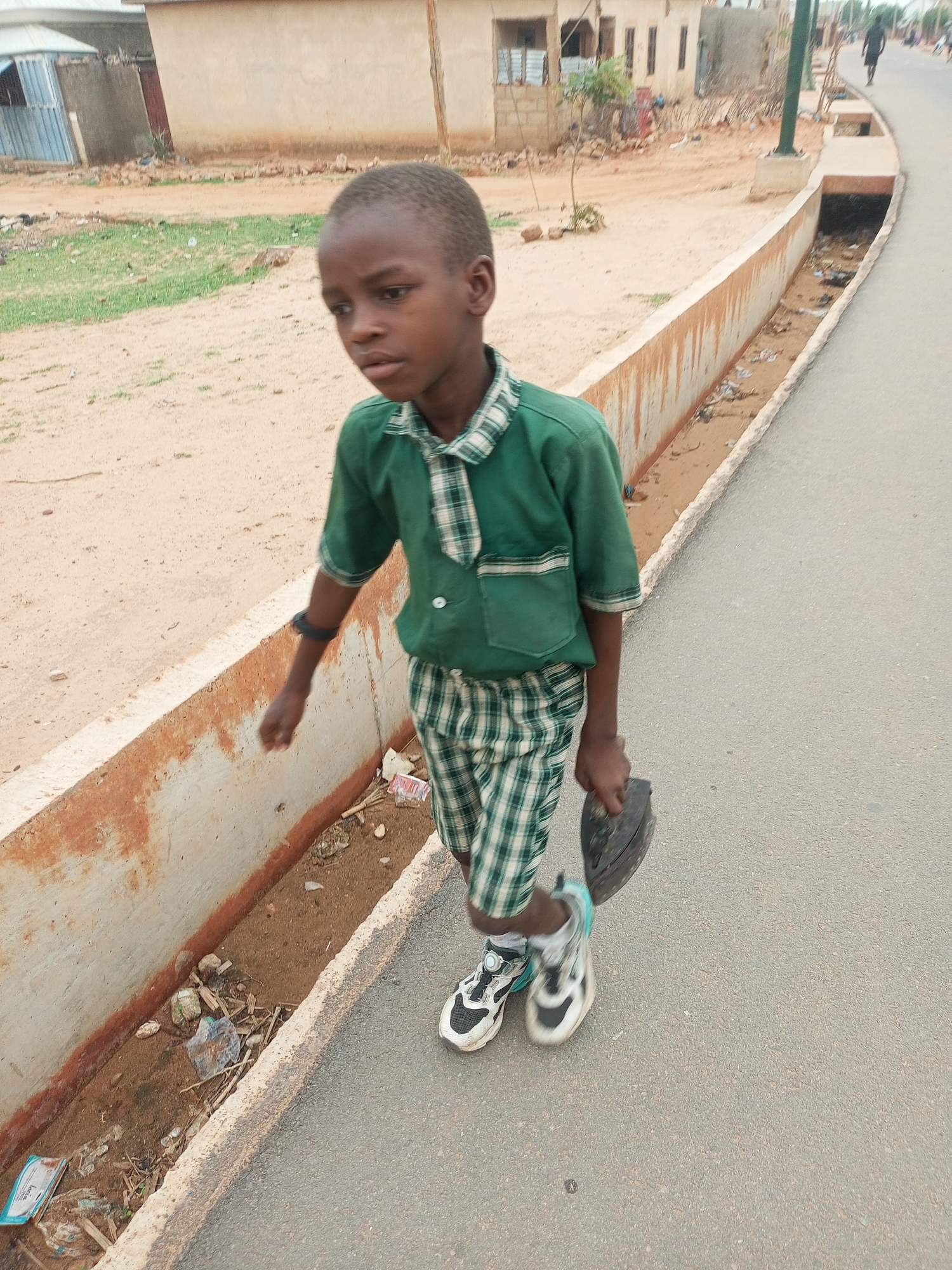 A young boy wearing a green and white plaid school uniform walks along the side of a paved road in Tempure, Nigeria. He carries a heavy, possibly antique, iron in his right hand. He is wearing sneakers and has a black band on his left wrist. The boy walks next to a concrete drainage ditch filled with trash and debris. A dirt road runs parallel to the paved road, with buildings visible in the background. Another person walks in the distance, further up the paved road.