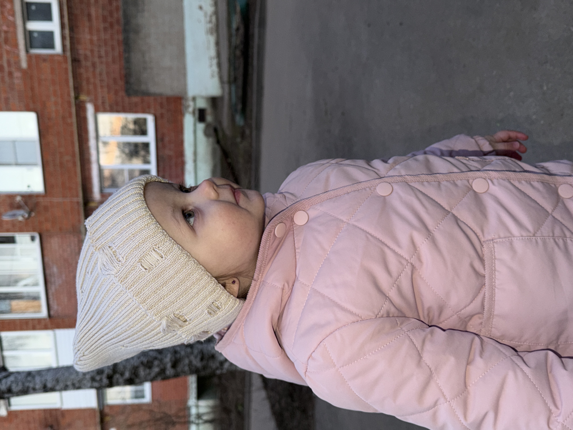 A young child wearing a light pink quilted jacket and a cream-colored knit hat is standing outdoors. The child is positioned in front of a brick building with several windows. The surroundings appear to be in the city of Reshetnikovo, Russia, based on geolocation. The background includes a concrete surface and some vegetation. The child is looking upwards and to the left.