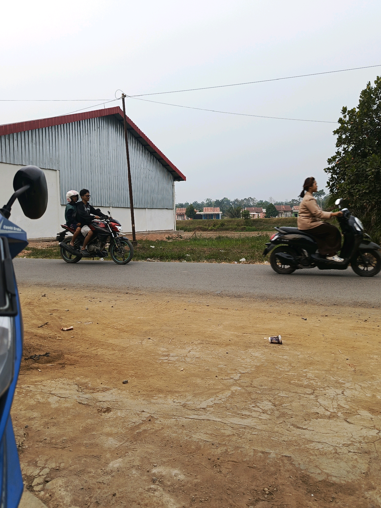 In Megang Sakti I, Indonesia, a scene unfolds on a paved road under an overcast sky. The foreground is dominated by a dirt and cracked concrete surface, with a blue object, possibly part of a scooter, visible on the left. A disposable cup lies on the ground in the mid-ground. In the middle distance, two individuals are riding on a red and black motorcycle, with the passenger wearing a pink helmet. To the right, a woman in a tan shirt and dark pants rides a black scooter. Behind them, a corrugated metal building with a red roof stands, and beyond that, a row of houses and vegetation suggest a residential area. The overall impression is of a quiet, possibly rural or suburban, setting.