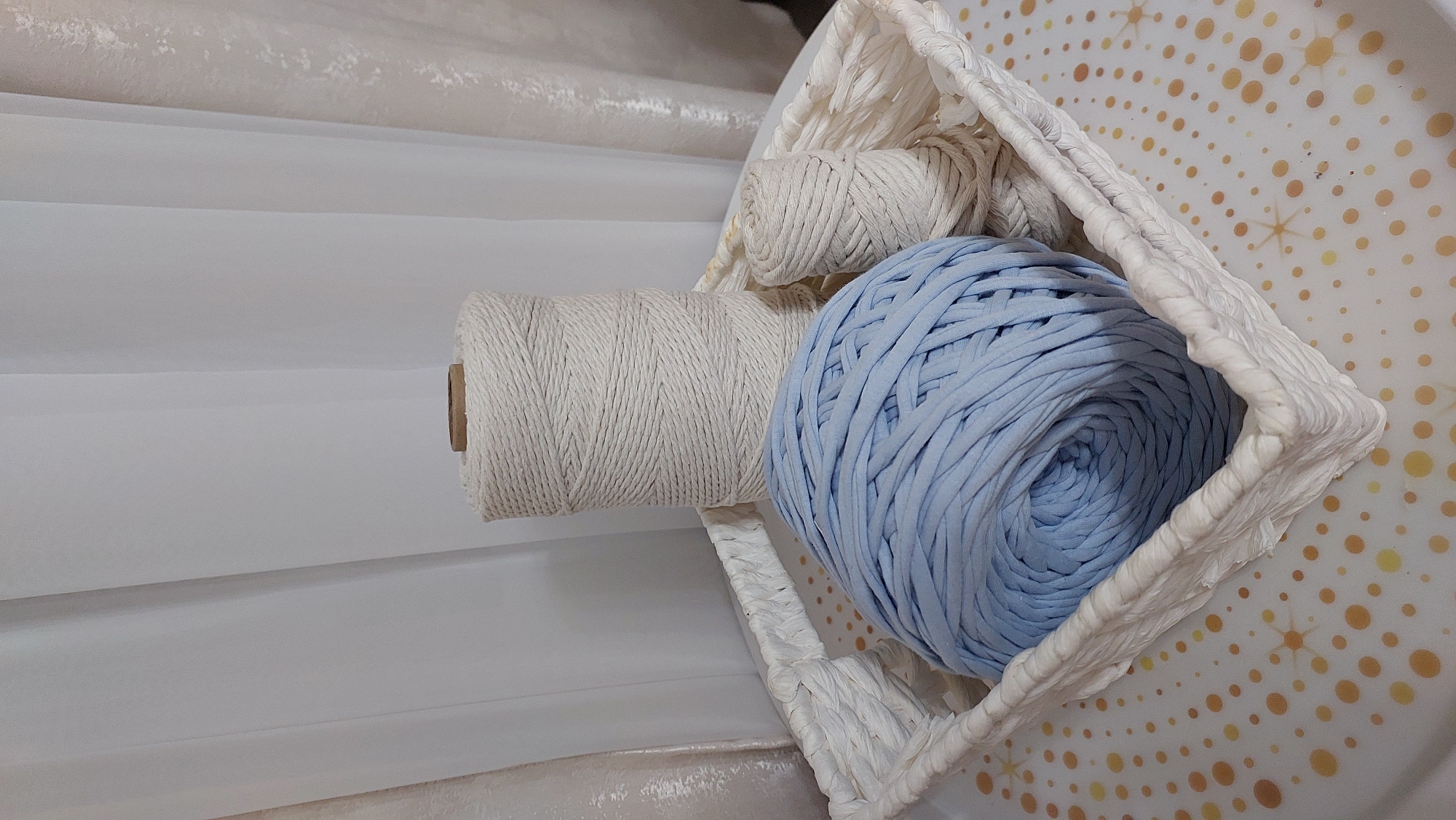 A white woven basket contains three spools of yarn. The largest spool is pale blue and tightly wound. To the right of the blue spool are two white spools of yarn, one larger than the other. The basket sits on a surface with a repeating pattern of gold starbursts and orange circles against a white background. Behind the basket, white sheer curtains are visible. The image is a still life, with no people or actions depicted. The location is not discernible from the image.