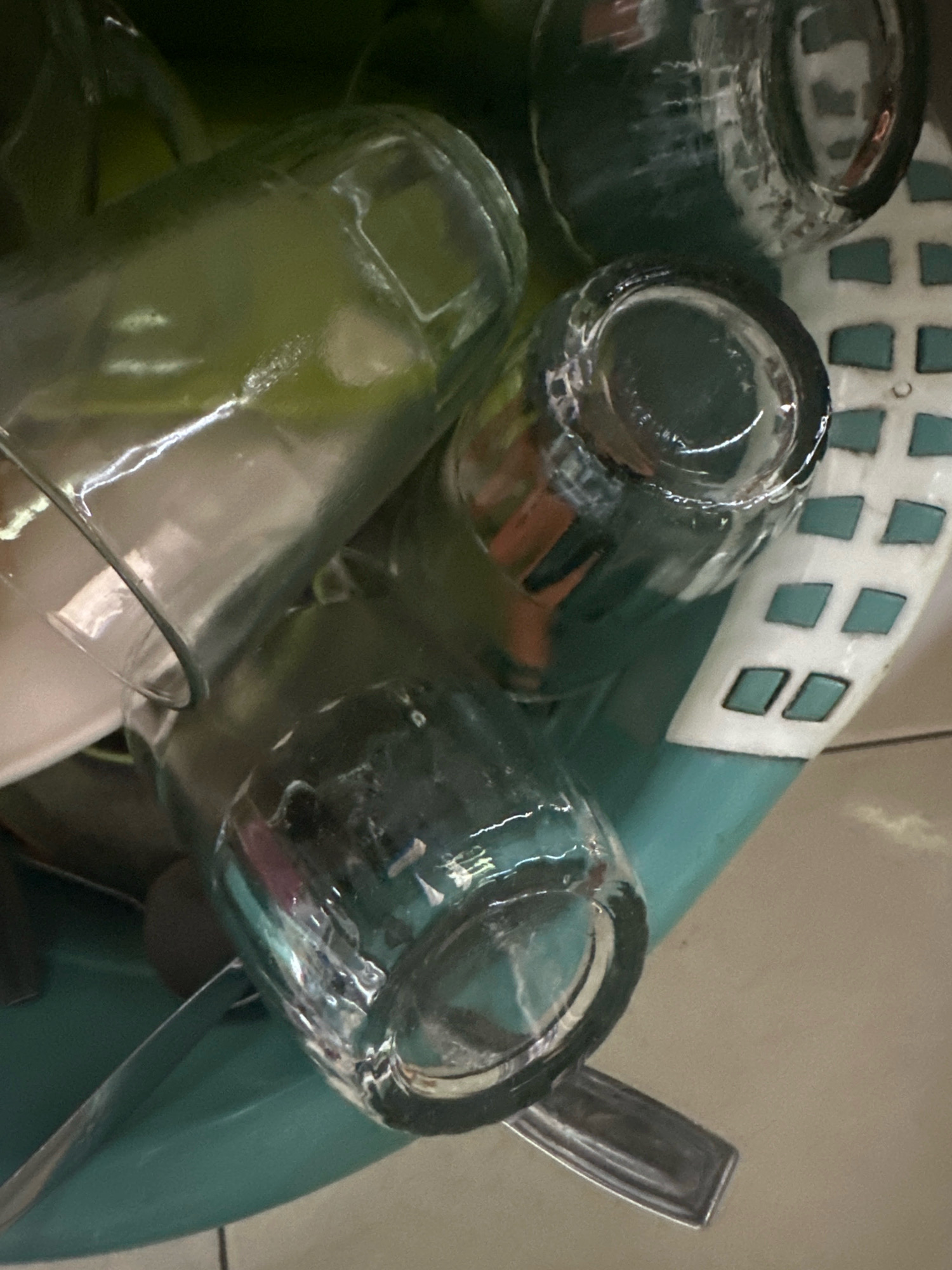 The image shows a close-up view of several clear glass jars and cups, some appearing to be dirty and stacked together in a teal-colored sink. The jars and cups are made of clear glass and have varying shapes and sizes. Some of the jars have a greenish tint, suggesting they might contain a liquid or residue. The sink appears to be made of plastic or ceramic. The background is a mixture of dark colors and some patterned white material, possibly a dish rack or part of the sink assembly. The lighting is dim, creating reflections and shadows on the glass surfaces. There are no people visible in the image. This scene is likely from a residential or commercial kitchen, possibly in Margamulya, Indonesia, given the context provided.