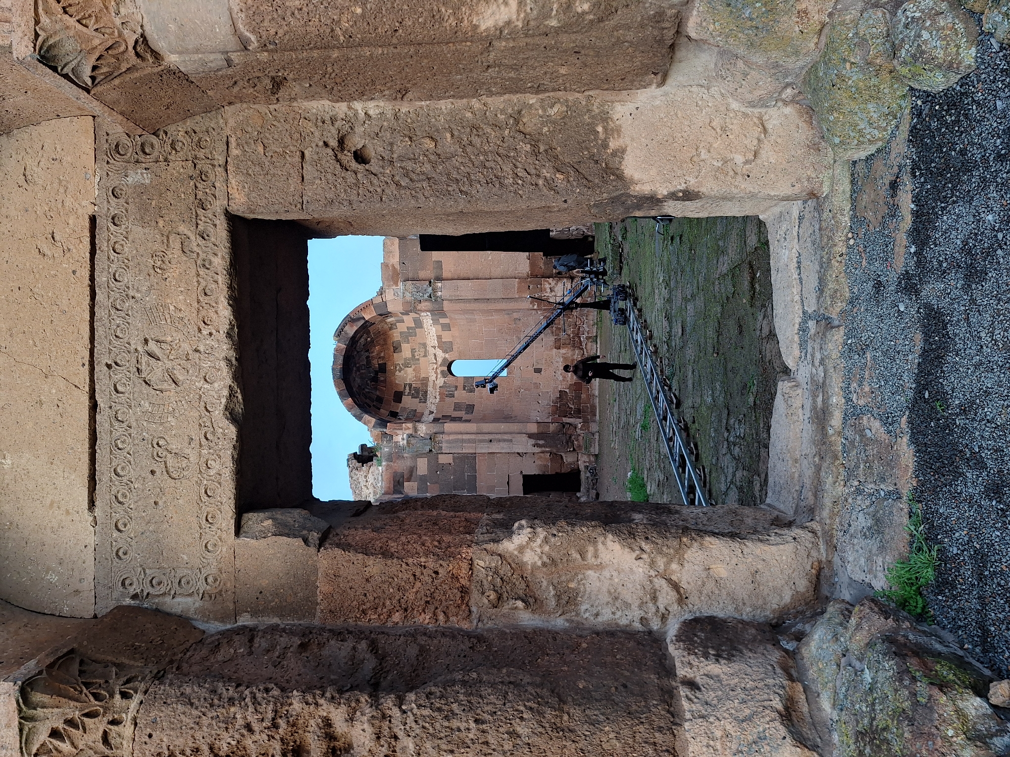 A low-angle, medium shot presents an architectural view through a stone-framed opening of an ancient structure, likely a church or similar building, located in Anipemza, Armenia. The viewer is looking through the stone opening towards the ruins. The frame's stonework on the left side is adorned with detailed carvings, with additional stone textures visible throughout the frame.
Outside the opening, a ladder and what appears to be film or recording equipment, as well as a human figure in dark attire, are visible. The ground is covered with a mixture of grass, gravel, and other vegetation. The background reveals the clear blue sky.