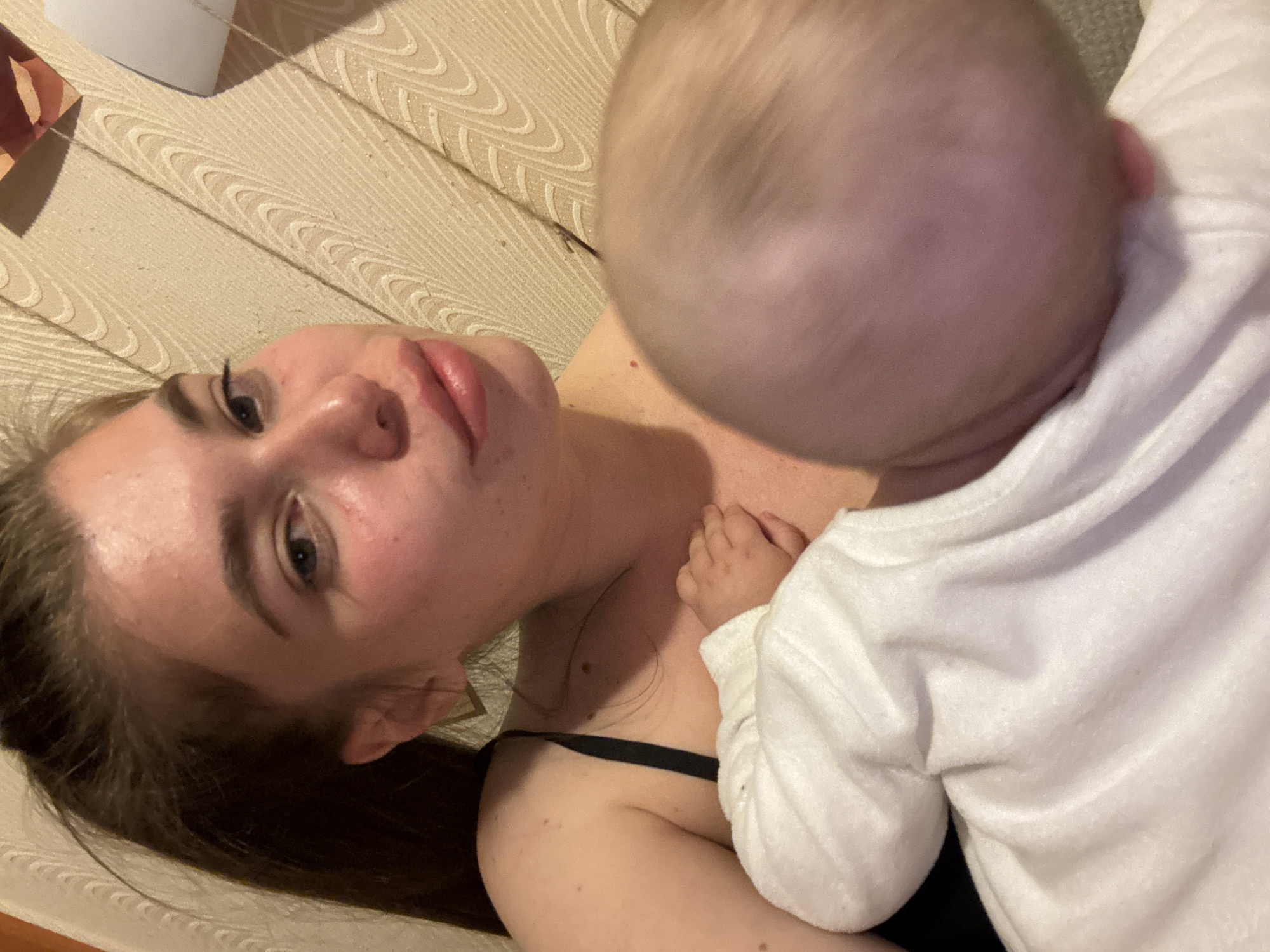 A woman lies on a textured, beige surface (possibly a bed or floor) with a baby lying on her chest. The woman has light skin, dark hair pulled back in a ponytail, and is wearing a black top. The baby has fair skin and light hair, and is wearing a white shirt. The baby's hand is resting on the woman's chest. The location in the city of Meshchery, Russia is unknown and not evident in the image.
