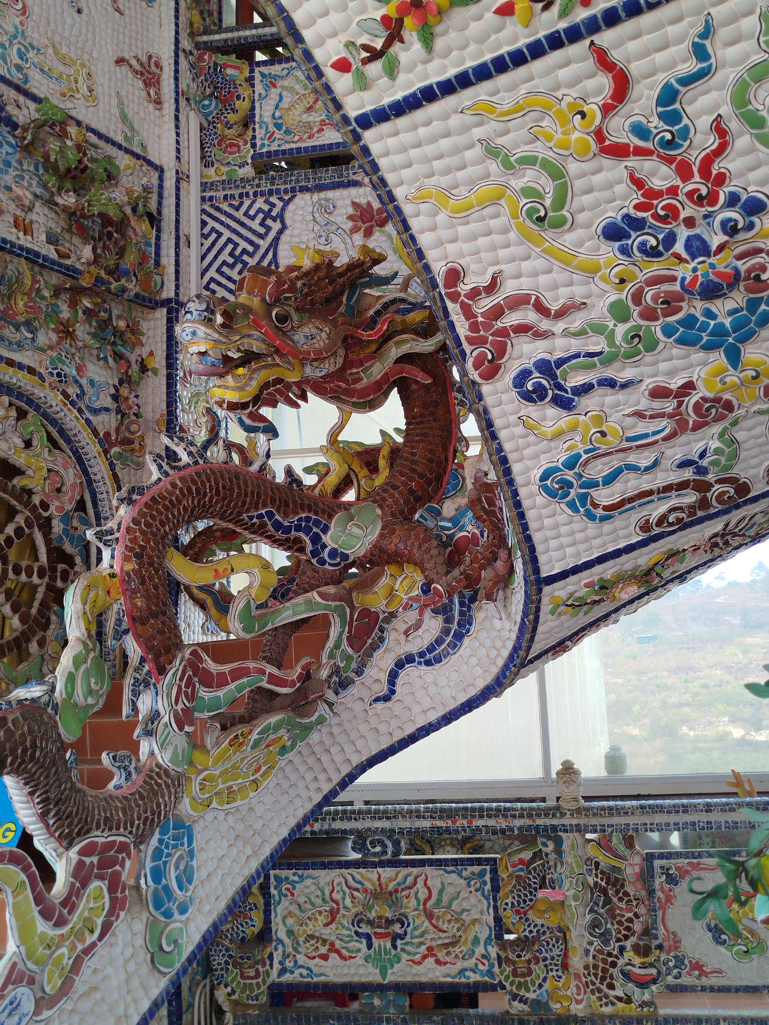 A detailed view showcases the intricate mosaic art of the Linh Phuoc Pagoda, also known as the Ve Chai Pagoda, located in Xuân Thành, Vietnam. The artwork features a large, colorful mosaic dragon snaking around an architectural structure. The dragon is predominantly red and brown, with accents of blue, green, and yellow scales, its body winding around an edifice covered in tiny white mosaic tiles. The background behind the dragon shows a window with a view of a distant hill or mountain.
The building itself is heavily decorated with colorful mosaic designs. The ceiling features patterns of clouds in white, blue, red, yellow, and green. The surface is also made up of the small white tiles. Below the dragon, more mosaic panels are visible, depicting floral designs and other dragons. The walls and surfaces are densely packed with this mosaic art, giving the entire scene a rich, ornate appearance.