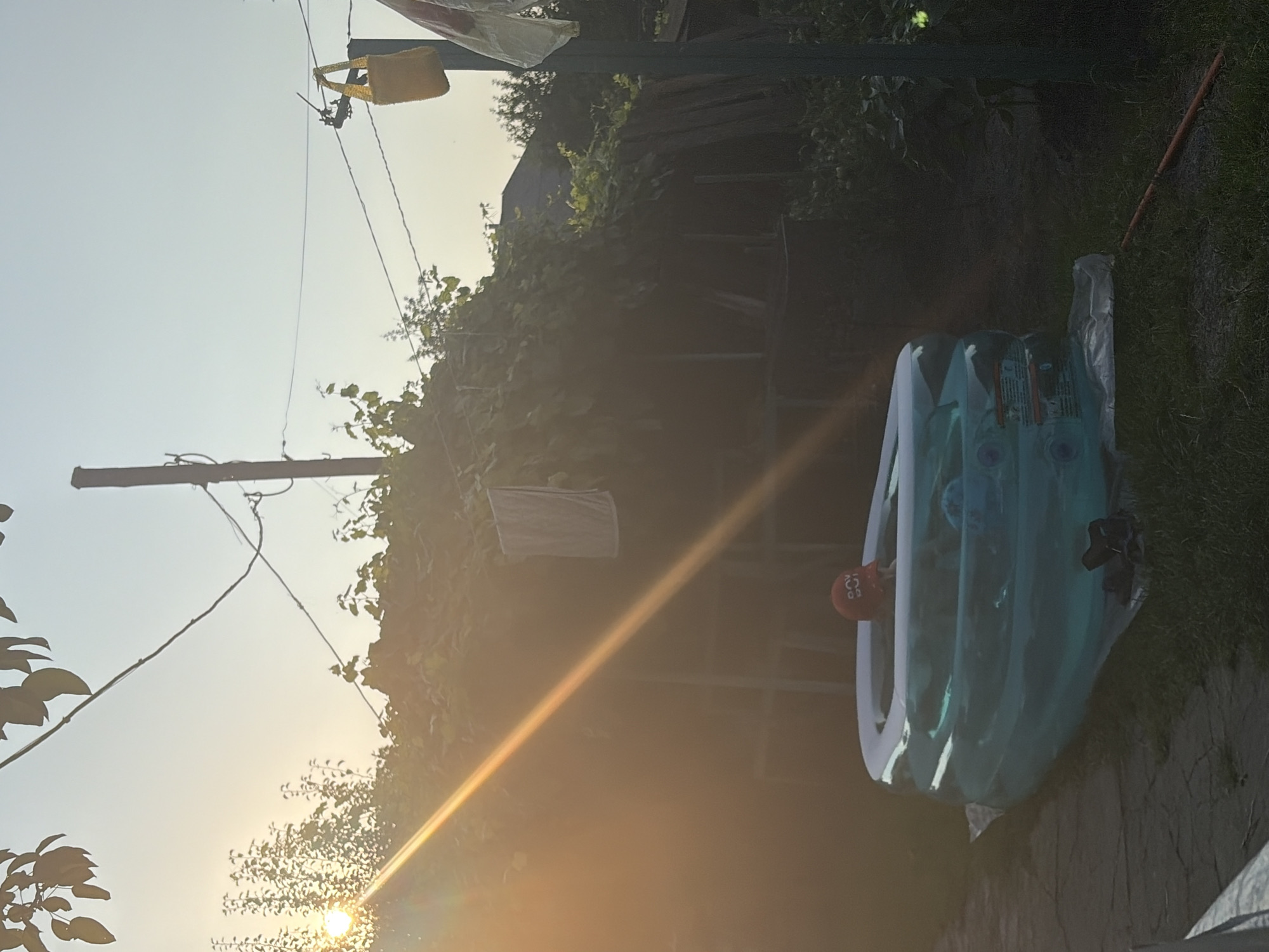 A child wearing a red hat with white lettering is sitting in a partially filled, blue inflatable kiddie pool in a backyard. The pool is placed on a patch of grass. The sun is setting, casting a warm glow and long shadows across the scene. Overhead, power lines crisscross the sky, one supporting a yellow bag and another with a white cloth hanging from it. A wooden pole stands prominently in the background, with vegetation and the side of a building visible further back. The ground is a mix of grass and a paved walkway.