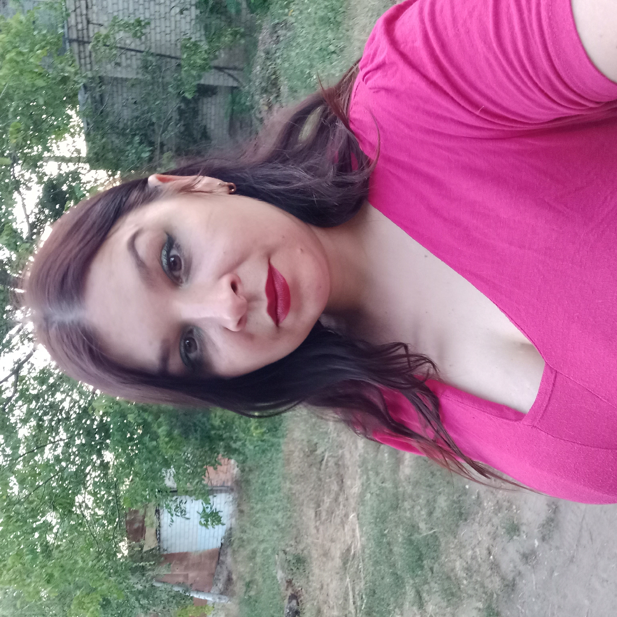 A woman with dark hair and green eyeshadow poses for a selfie outdoors. She is wearing a bright pink top with a V-neck. In the background, there are trees, a brick building, and dry grass. The location is identified as Unknown, Ukraine.