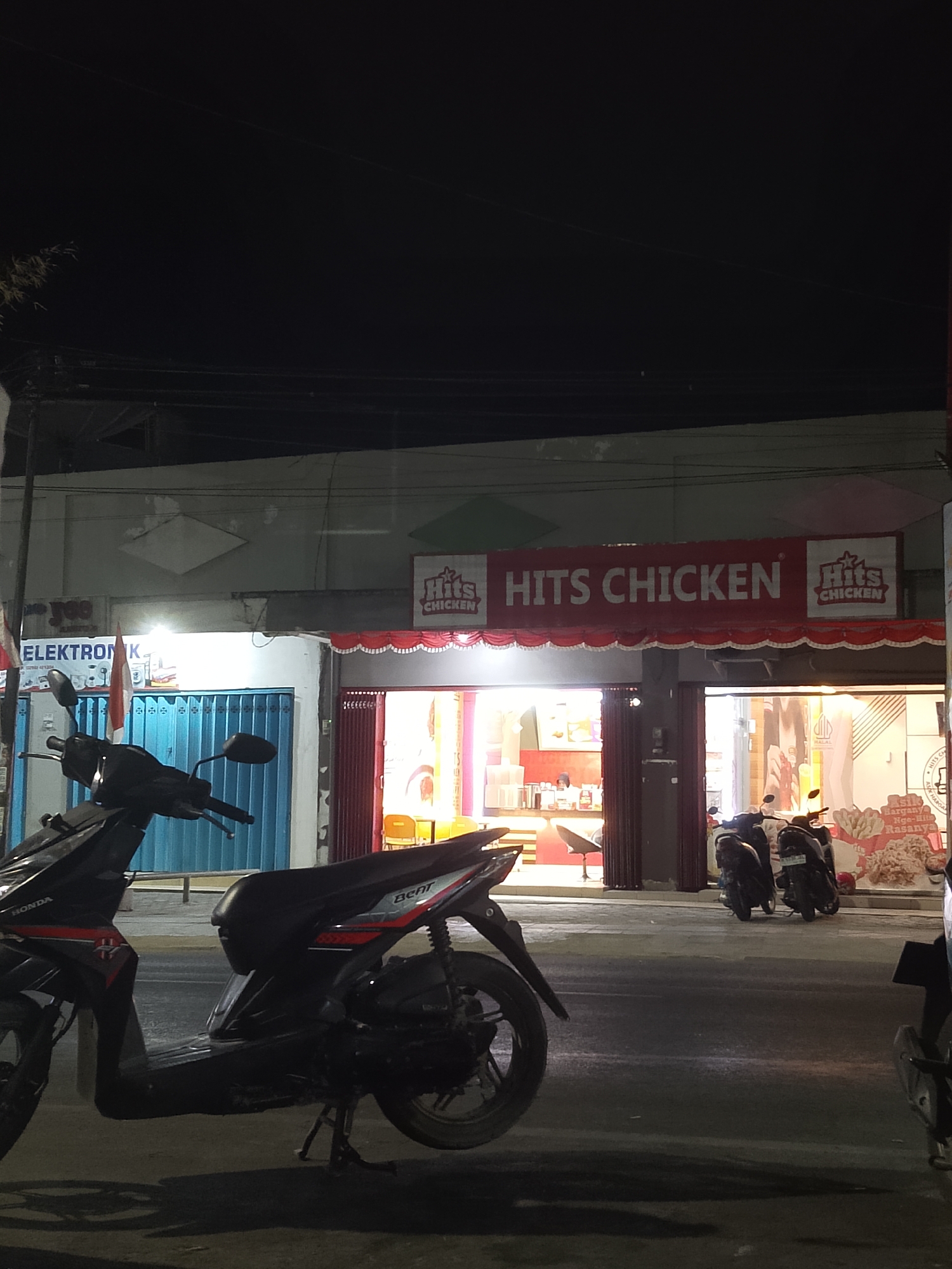 The image depicts a nighttime street scene in Balun, Indonesia, likely a commercial area. A brightly lit storefront with a red awning and prominent signage reading "HITS CHICKEN" is the central focus. The storefront appears to be a fast-food establishment, with interior lights illuminating displays and seating areas. In front of the "HITS CHICKEN" establishment, several motorcycles are parked along the street.
To the left of the "HITS CHICKEN" store, another establishment is visible with a blue shuttered entrance and signage that partially reads "ELEKTRONIK". An Indonesian flag is displayed next to this store. The street is paved and appears to be in moderate condition. The lighting is artificial, casting shadows from the motorcycles and the buildings onto the road. The sky is dark, indicating nighttime. There are no visible people in the immediate foreground, but figures might be present inside the lit establishments. The overall impression is that of a typical urban commercial street at night.