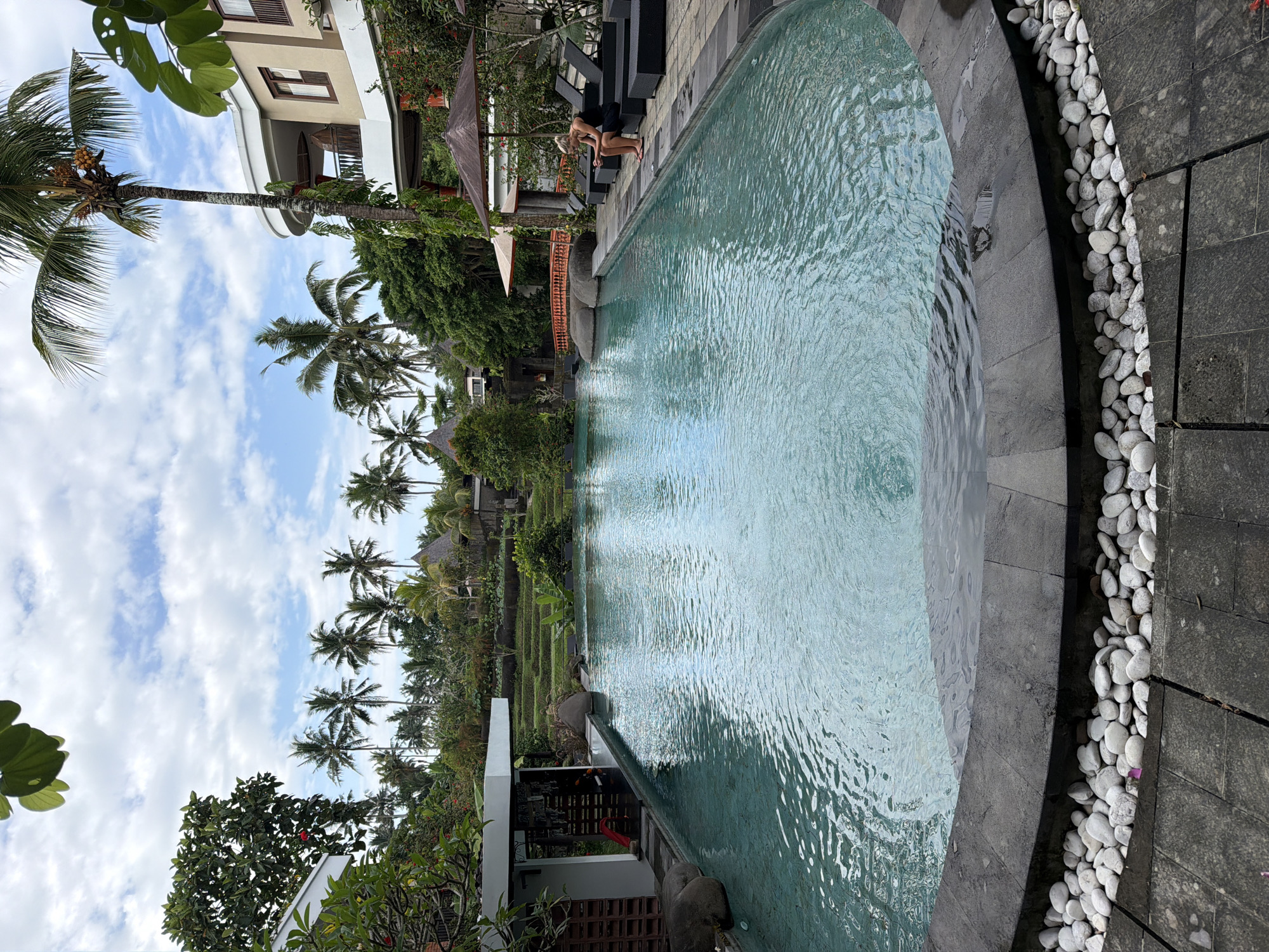 The scene depicts a serene, outdoor swimming pool at a location in Peliatan, Indonesia. The pool's edge is curved, lined with gray stones and accented by a bed of white pebbles. A woman is seated on a dark lounge chair next to the pool. Palm trees tower in the background, adding to the tropical ambiance, set against a backdrop of a blue sky with scattered clouds. Lush greenery surrounds the area, with a building and possible rice terraces visible in the distance. A covered structure is also situated nearby, adding architectural detail to the setting.