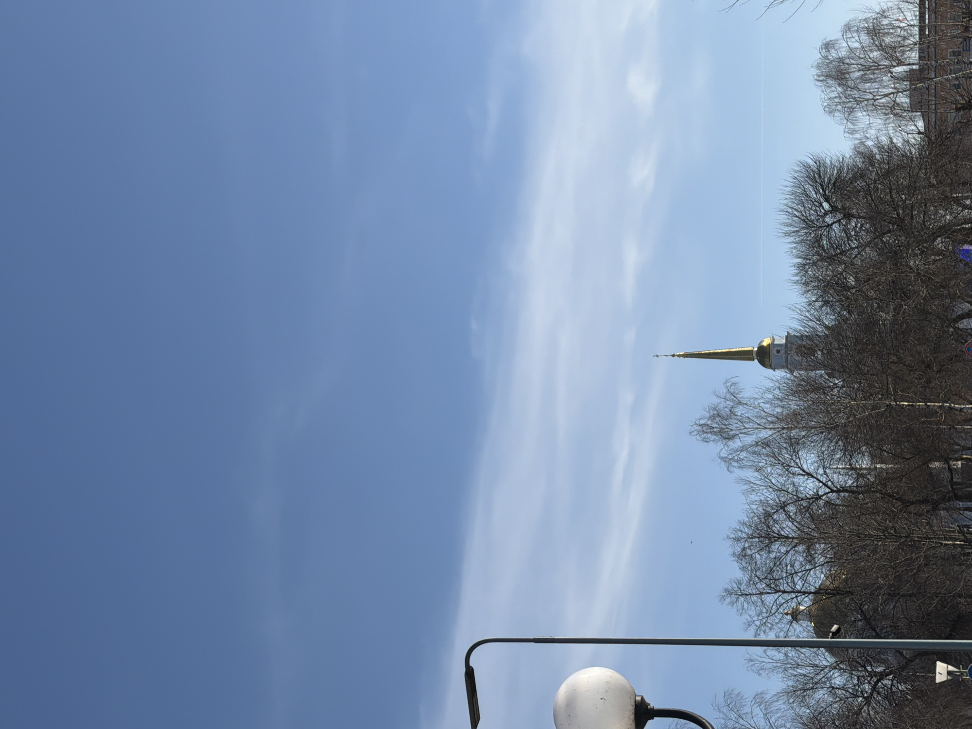 A ground-level, upward-facing shot shows a clear blue sky with elongated wispy clouds, partially obscured by the bare branches of trees. A gold-topped spire extends above the trees. In the foreground, a streetlight fixture is visible. This location is Votkinsk, Russia.