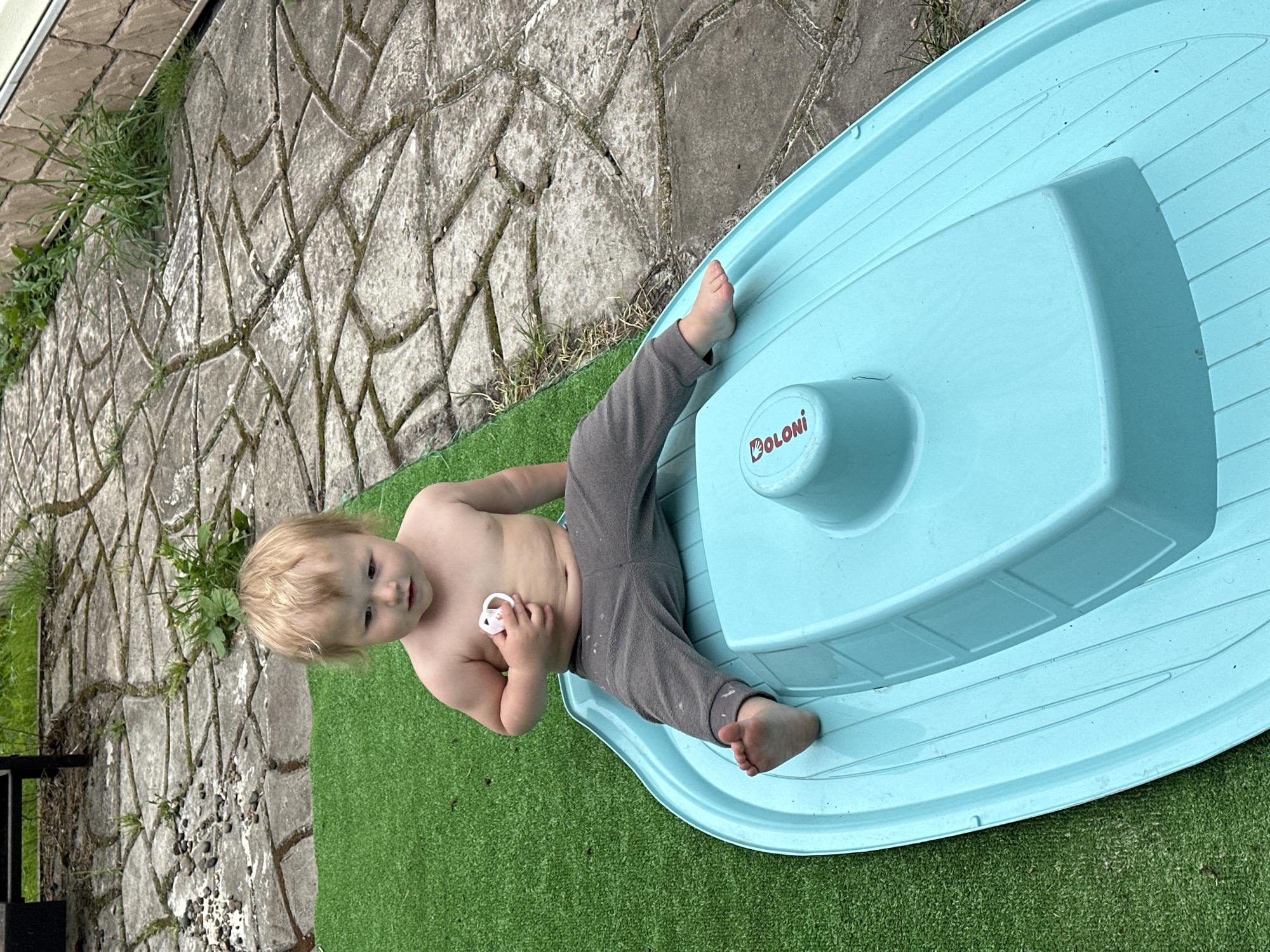 A shirtless toddler with blonde hair sits on a blue plastic toy boat on a patch of artificial grass. The toddler is wearing grey pants and holding a white pacifier in their right hand. The boat has the word "COLONI" printed on it in red. The artificial grass is next to a paved area made of irregular concrete stones with some weeds growing between them. In the background, there is a wall made of light brown bricks and some greenery. The scene appears to be an outdoor residential area. The location is not specified in the image.