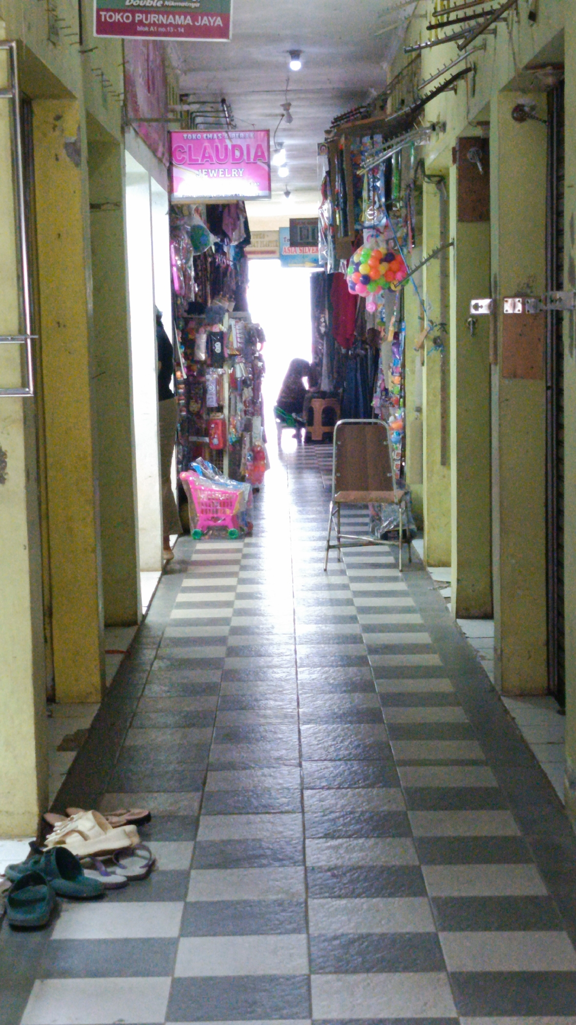 The image depicts a narrow indoor walkway or corridor, possibly a market or a row of shops, in Kertajaya, Indonesia. The walkway is paved with alternating dark and light checkered tiles. On the left, there are shops with yellow-painted walls and metal shelving. A prominent pink sign that reads "TOKO PURNAMA JAYA" is visible at the top of the left wall. Further down, another pink neon sign displays "CLAUDIA JEWELRY." Various items are displayed for sale on racks and shelves along both sides of the corridor, including clothing, accessories, and colorful decorative items. A small pink plastic toy shopping cart is situated in the middle of the walkway on the left. Further down the corridor, a woman wearing dark clothing is seated on a stool in the dim light, facing away from the camera, near a stall displaying goods. On the right side of the walkway, a brown metal chair with a wooden seat and back is positioned. Several pairs of flip-flops and shoes are scattered on the floor in the foreground on the left side of the image. The ceiling is a functional, industrial-looking structure with exposed pipes and lighting fixtures. The overall impression is a bustling commercial space, albeit with areas of shadow and a slightly cluttered appearance.