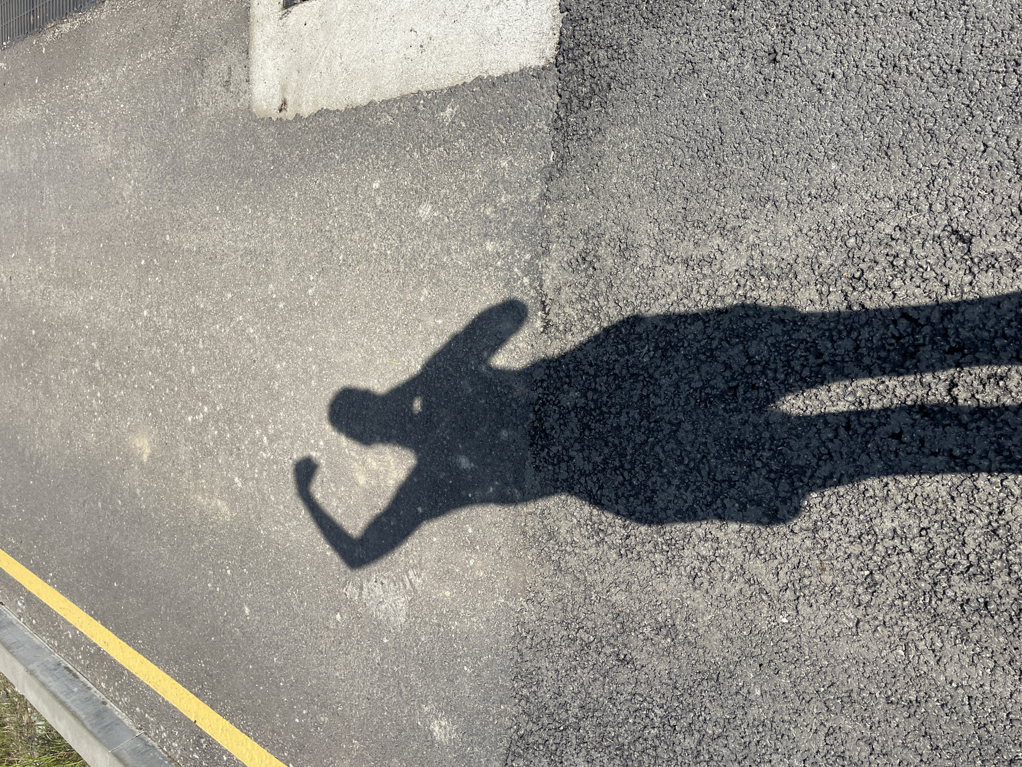 The image shows a shadow of a person on an asphalt surface, possibly a street or pavement. The shadow suggests the person is standing and raising one arm. A yellow line is visible near the bottom left corner, indicating a road marking. The asphalt surface has varying textures and appears to be two different types of pavement. A concrete curb is also visible on the left side. No specific details in the image point to Sochi, Russia.