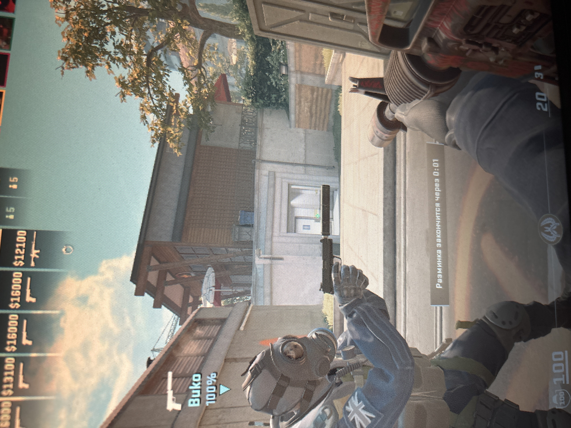 The image is a first-person perspective from a video game, likely Counter-Strike: Global Offensive, set in an urban environment. In the foreground, the player's gloved hands hold a weapon, partially visible. A character in a gas mask and tactical gear, identified as "Buko" with 100% health, is visible to the left, holding a handgun. In the background, a two-story building with a modern architectural style is prominent, featuring wooden panels and large windows. Trees with autumn foliage are present, and the sky is a pale blue with scattered clouds. A notification in Russian, "Разминка закончится через 0:01," indicating "Warm-up will end in 0:01," is displayed. The overall scene depicts a moment of preparation or transition within a game match.