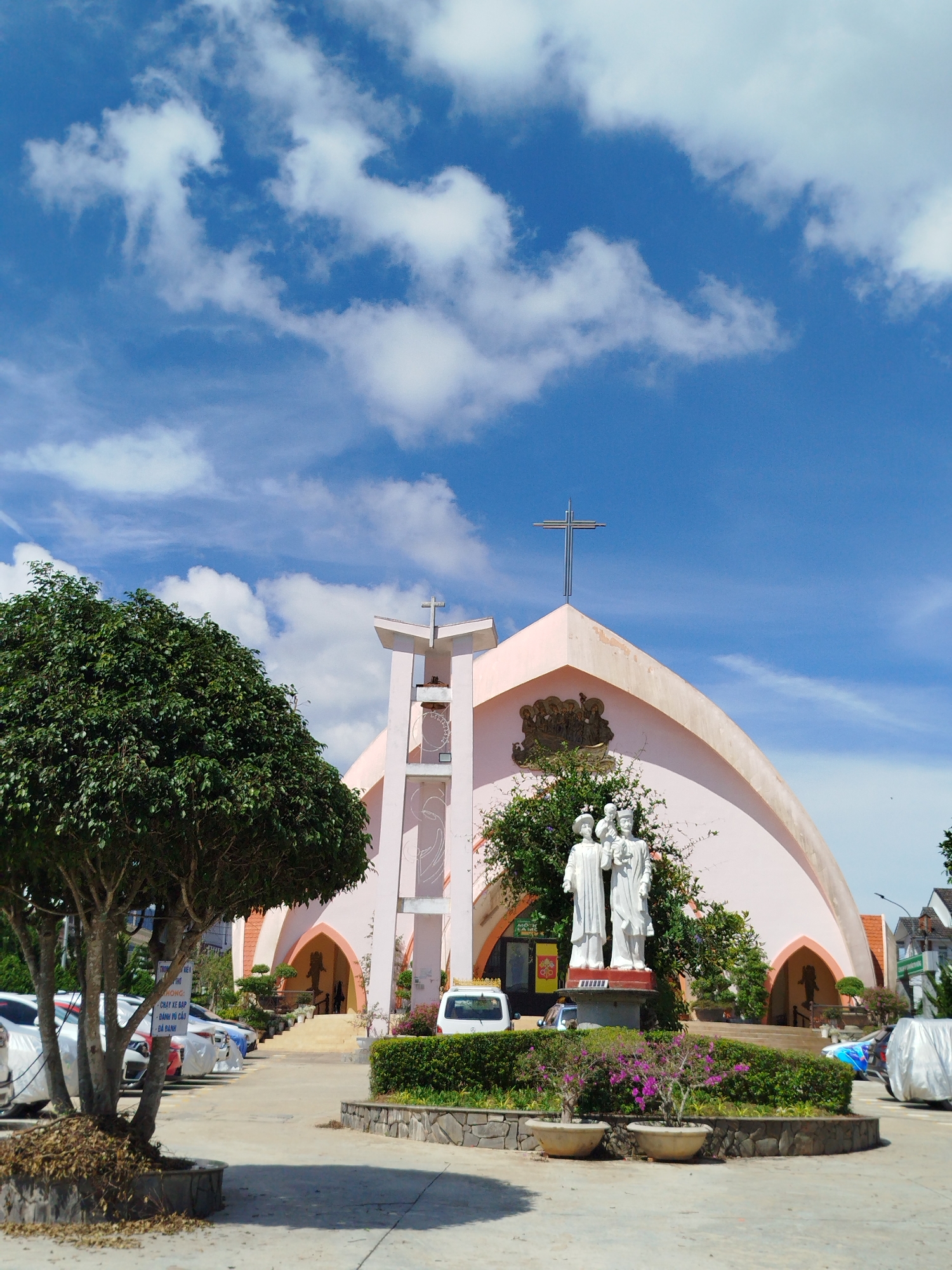 The image shows a pale pink church with a modern, arched design, topped with a metal cross. A tall, thin bell tower stands to the left of the church. In front of the church is a statue of the Holy Family (Jesus, Mary, and Joseph), surrounded by greenery. A tree on the left obscures some of the parking area, where several cars are parked. The sky is bright blue with scattered white clouds. The image shows the location in the city of Cam Ly - Đà Lạt, Vietnam. The church is well maintained, with manicured landscaping and clear signage. A paved area in the foreground leads to the church entrance.