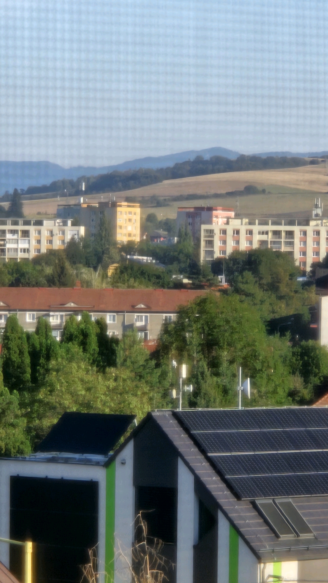 The image shows a view of a residential area in Prešov, Slovakia, with buildings and vegetation. In the foreground, two modern structures with angled roofs are visible, equipped with numerous dark solar panels. One structure has a prominent green vertical stripe. Behind these, a row of apartment buildings with red tile roofs is present, interspersed with trees. Further in the background, taller apartment buildings, some with light-colored facades and balconies, and one with a bright yellow facade, are visible. Beyond the buildings, rolling hills covered in fields and trees form the landscape, under a clear, pale blue sky. The overall impression is a suburban or semi-urban environment during daylight.