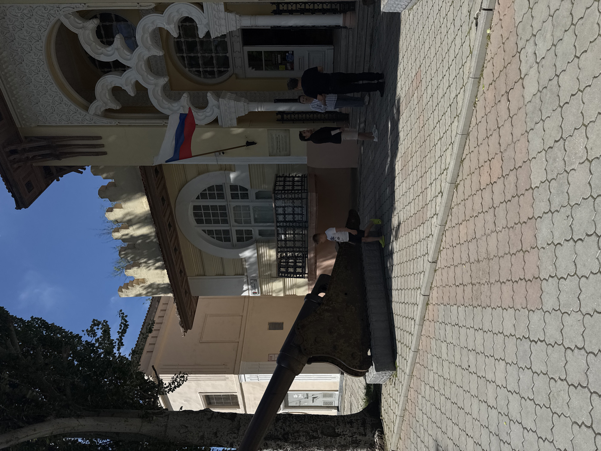 The image depicts a street scene in Yevpatoriya, Ukraine. A Russian flag is prominently displayed on the building. Three individuals are standing near the entrance of the building. A boy is near a canon that is positioned on a low stone base. The architecture features decorative elements, including ornate window frames and a balcony with a metal railing. The pavement is made of interlocking stones, and a tree partially obscures the view of the lower part of the building.