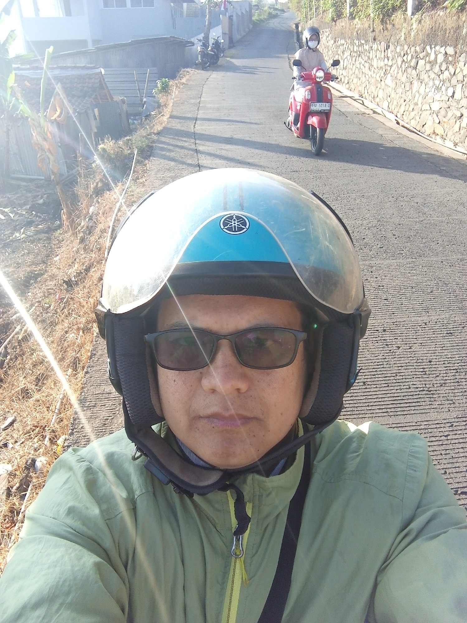 A POV shot from a motorcycle helmet captures a person wearing a light green jacket and sunglasses, with a blue helmet. In the background, another person on a red scooter drives away down a paved road. The road is lined with a stone wall on the right and dry vegetation and small structures on the left. The scene appears to be in a semi-rural area, possibly Cihanjuang, Indonesia, given the architecture and vegetation. The lighting suggests it is daytime, with the sun casting strong shadows and lens flares.