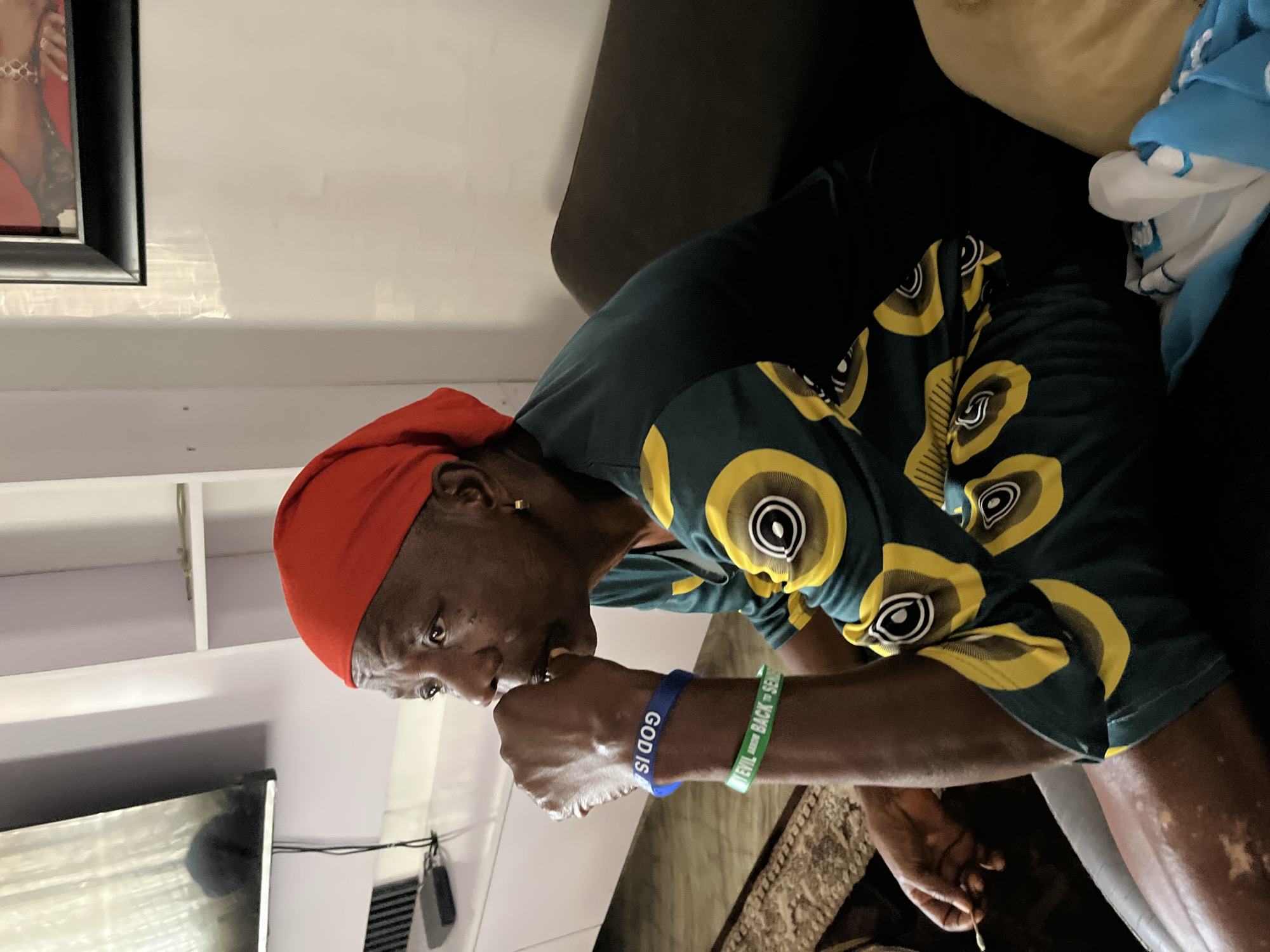A vertically oriented photograph, taken indoors, depicts an older individual seated on a sofa in what appears to be a living room in Jos, Nigeria. The subject, who has dark skin and noticeable skin discoloration (possibly vitiligo) on their left leg and hand, is seen from the waist up. They are wearing a red headscarf and a dark green shirt with a distinctive yellow and black circular pattern. A single gold earring is visible on their right ear.
The person is turned to their left, with their gaze directed downwards. Their right hand is raised towards their mouth, possibly holding something small or in a pensive gesture. On their left wrist, they wear two silicone wristbands: a blue one with the visible text "GOD IS" and a green one with partially discernible text, appearing to say "...EVIL...BACK = SEND".
In the background, elements of the room are visible, including a dark-framed picture on a white wall, a white shelving unit, and a dark sofa. A rug is visible on the floor. The lighting suggests a bright indoor environment, likely during the day.