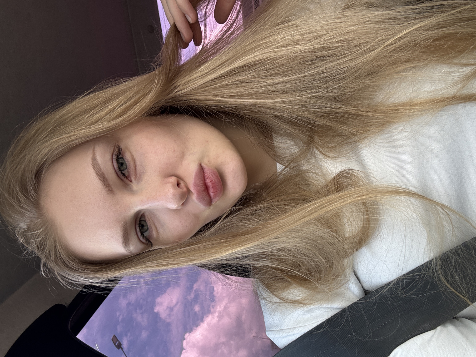 A close-up, slightly tilted portrait features a young woman with long, straight blonde hair flowing around her. Her face is prominent, showcasing light-colored eyes (appearing greenish-grey) with visible eyelashes and well-groomed eyebrows. Her full lips are slightly parted, revealing a natural pink hue. She has fair skin and is wearing a light-colored top, likely white.
The woman appears to be inside a vehicle, as indicated by a dark grey seatbelt visible across the lower right of the frame. Her hand is partially visible at the top of the image, seemingly adjusting her hair. Through a window on the left, a striking sky with vibrant pink and purple clouds is visible, suggesting either sunrise or sunset. A dark silhouette of a lamp post is subtly visible against the colorful sky in the bottom left corner, hinting at an urban or roadside environment. Her expression is calm and neutral, looking directly towards the camera. The image's factual location is Perm, Russia. No text is visible in the media.