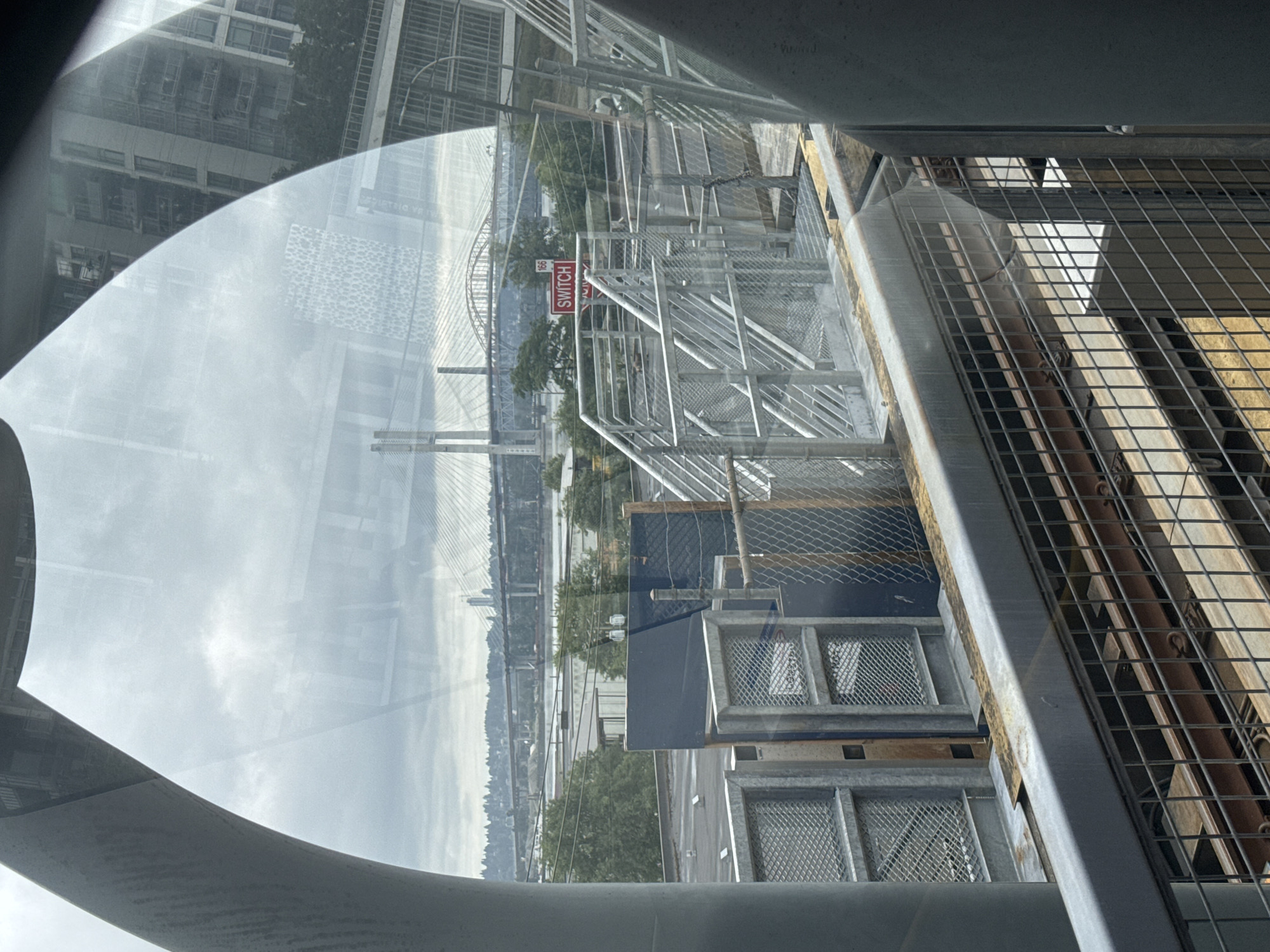The image captures a view from a transit vehicle, likely a SkyTrain, in New Westminster, Canada. A large, curved gray structure frames the left side of the scene, suggesting the interior of the transit car. Through the window, a cloudy sky looms over an urban and industrial landscape. In the distance, a massive suspension bridge with towering pylons spans a body of water, likely the Fraser River. Numerous other bridges and urban structures are visible along the horizon. Closer to the foreground, a network of metal stairs and railings suggests a SkyTrain station or an elevated walkway. Several industrial buildings, some with dark blue siding, are present. A red sign with the word "SWITCH" is visible on a pole, along with a number "166". The scene also includes chain-link fences, metal grates, and what appear to be train tracks below the elevated structure. The overall impression is one of a functional, modern city with a strong emphasis on transportation infrastructure.