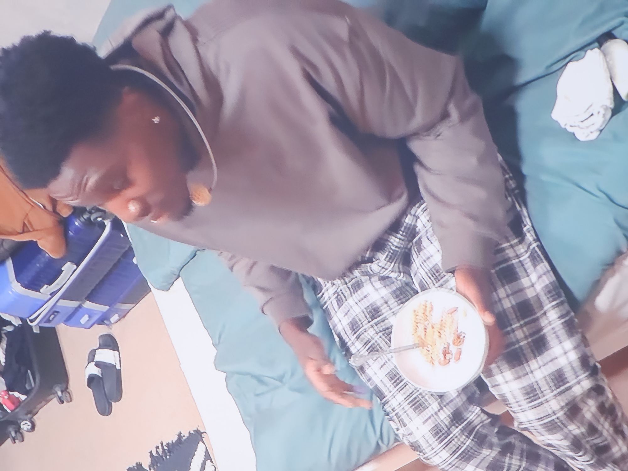 The image depicts a man sitting on a turquoise bed with a bowl of cereal in his hands. He is wearing a brown hoodie and plaid pants. The background shows luggage and a pair of flip-flops on the floor. The scene appears to be in a room. The exact location in the city of Buchen (Odenwald), Germany cannot be determined from the image alone.