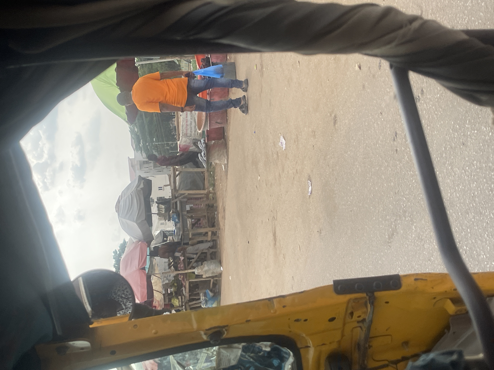 The image depicts an outdoor scene, likely a marketplace, in Galadimawa Village, Nigeria. In the foreground, a yellow vehicle, possibly a rickshaw or a similar motorized tricycle, is partially visible. The perspective suggests the image is taken from inside this vehicle. In the mid-ground, a market area is set up with wooden stalls and various goods displayed. Several individuals are present, some tending to the stalls and others browsing. One man, wearing an orange t-shirt and blue jeans, is walking away from the viewer, carrying a blue plastic bag. Umbrellas, one green and maroon, and another pink, are erected to provide shade. Buildings, seemingly residential, are visible in the background. The ground is paved with dirt and asphalt. The overall atmosphere suggests a typical day at a local Nigerian market.