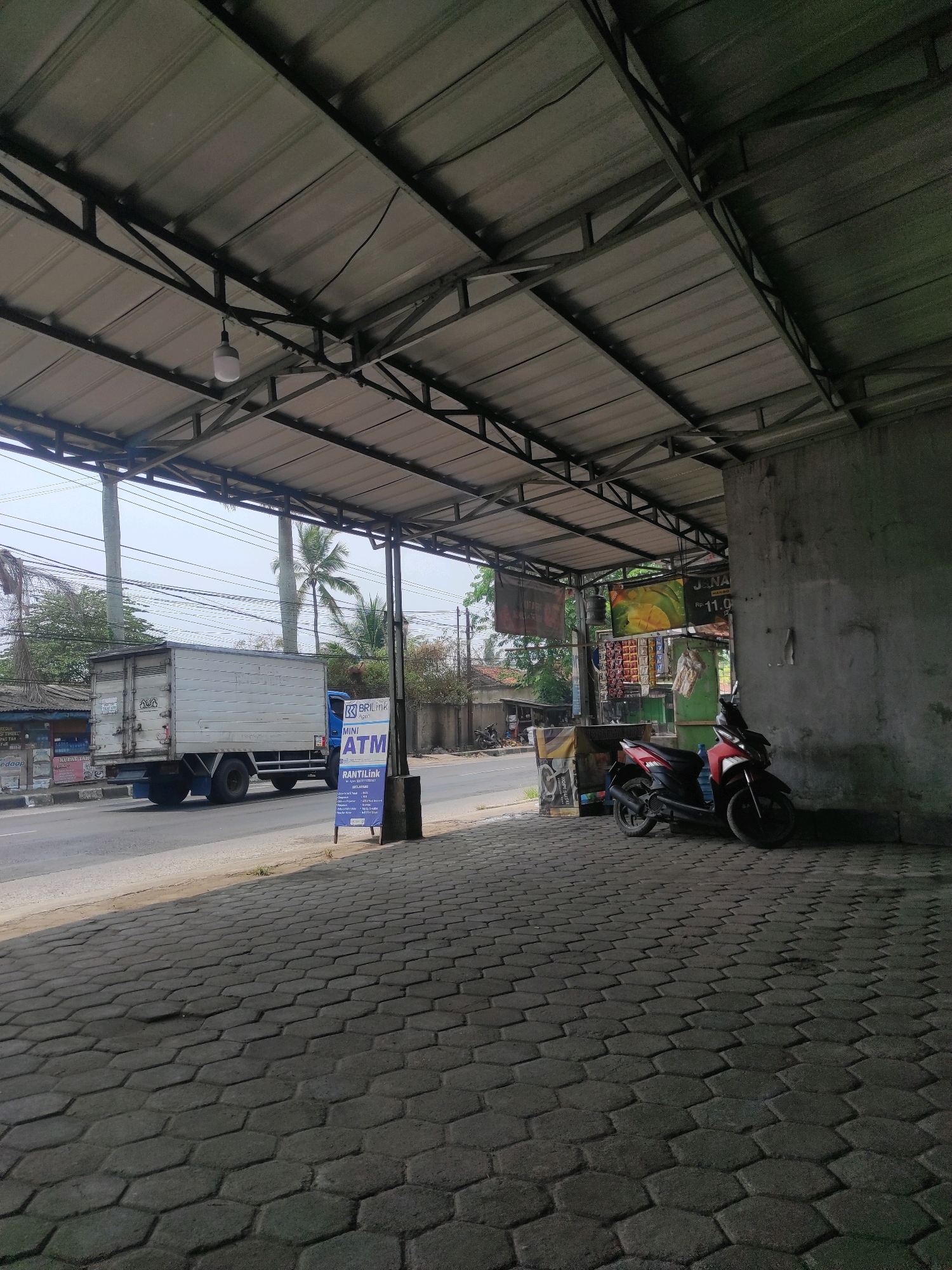 The image shows a street scene in Cipatat, Indonesia, under a corrugated metal awning supported by a metal frame. The awning extends over a paved walkway made of interlocking hexagonal pavers. To the left, a white box truck is driving on the road, which appears to be a two-lane asphalt road. A blue signpost with "ATM" and "BRI Link" branding stands near the edge of the walkway, in front of a concrete wall. Parked on the walkway, leaning against the concrete wall, is a red and black motorcycle. The background shows trees, including palm trees, utility poles with wires, and a few buildings. The overall impression is a commercial or service area along a road.