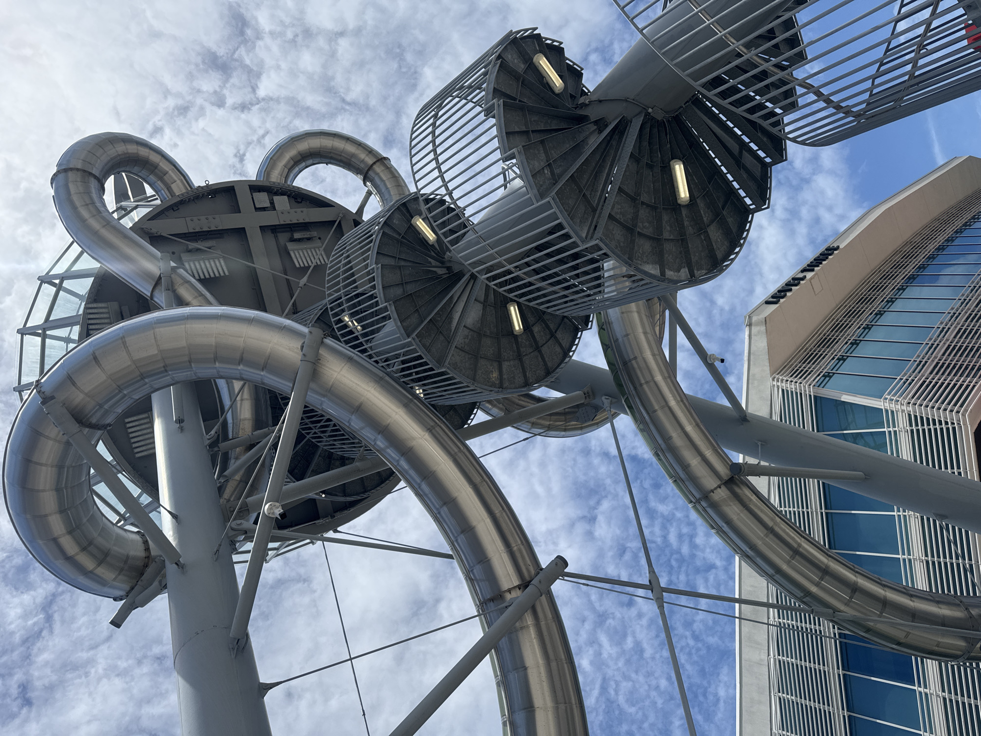 The image shows a low-angle, wide shot of a large, outdoor spiral slide structure in Aventura, Florida. The slide, made of shiny, segmented metal tubing, curves and twists upwards against a partly cloudy blue sky. Several grey, metal spiral staircases with railings are visible, attached to the slide's support structure. The staircases are illuminated by fluorescent lights. In the background, a modern building with blue windows and horizontal silver louvers is partially visible. The building has signage that reads "LEVEL THREE". The overall impression is of a large, complex, and modern amusement or attraction.