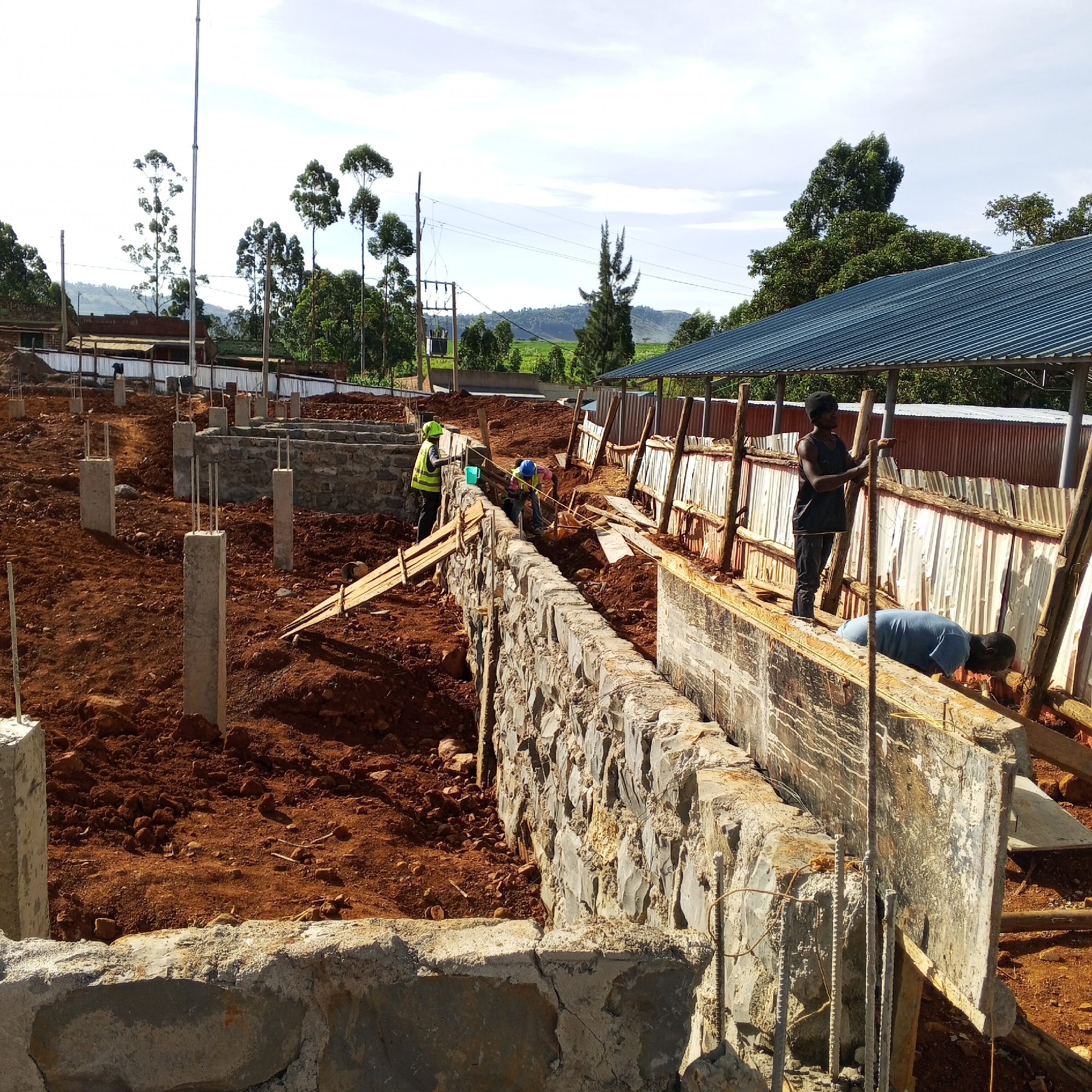 The image depicts a construction site in what is presumed to be Borabu Chitago ward, Kenya. Several concrete pillars are spaced across a reddish-brown dirt landscape, suggesting the foundation for a building. Workers are seen constructing a stone and concrete wall. Two workers in hardhats and safety vests are measuring the wall. Two other workers are inside the wall framework. The environment includes trees and power lines in the background. A structure with a blue corrugated metal roof and a corrugated metal wall is also visible in the background.