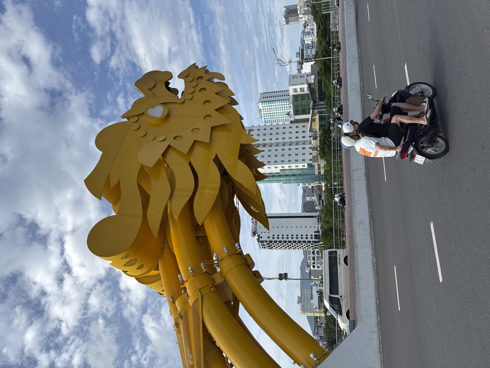A yellow dragon sculpture, part of the Dragon Bridge in Da Nang, Vietnam, dominates the frame against a partly cloudy sky. The sculpture's head and neck, made of segmented yellow tubes and plates, are prominently displayed on the left side of the image. In the background, modern city buildings rise, including a white tower with a distinctive black and white geometric pattern. A white van is parked on the right side of the bridge, and a man and woman are riding a dark-colored scooter on the road in the foreground. The road itself is asphalt, with white lane markings. The scene appears to be captured during daylight.