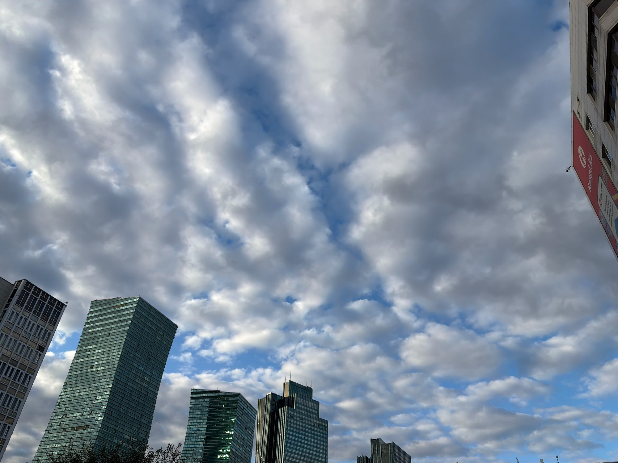 The image captures a view of the Astana skyline under a dramatic, cloudy sky. Several modern skyscrapers, characterized by their glass facades, dominate the lower portion of the frame. These buildings exhibit varying architectural styles, with some featuring distinct, leaning designs. A tall building on the far right of the image has a large red banner attached to its exterior, displaying white text. The sky is filled with a mix of dark gray and white clouds, with patches of blue sky visible in between. The overall impression is one of an urban landscape on a day with dynamic weather conditions.