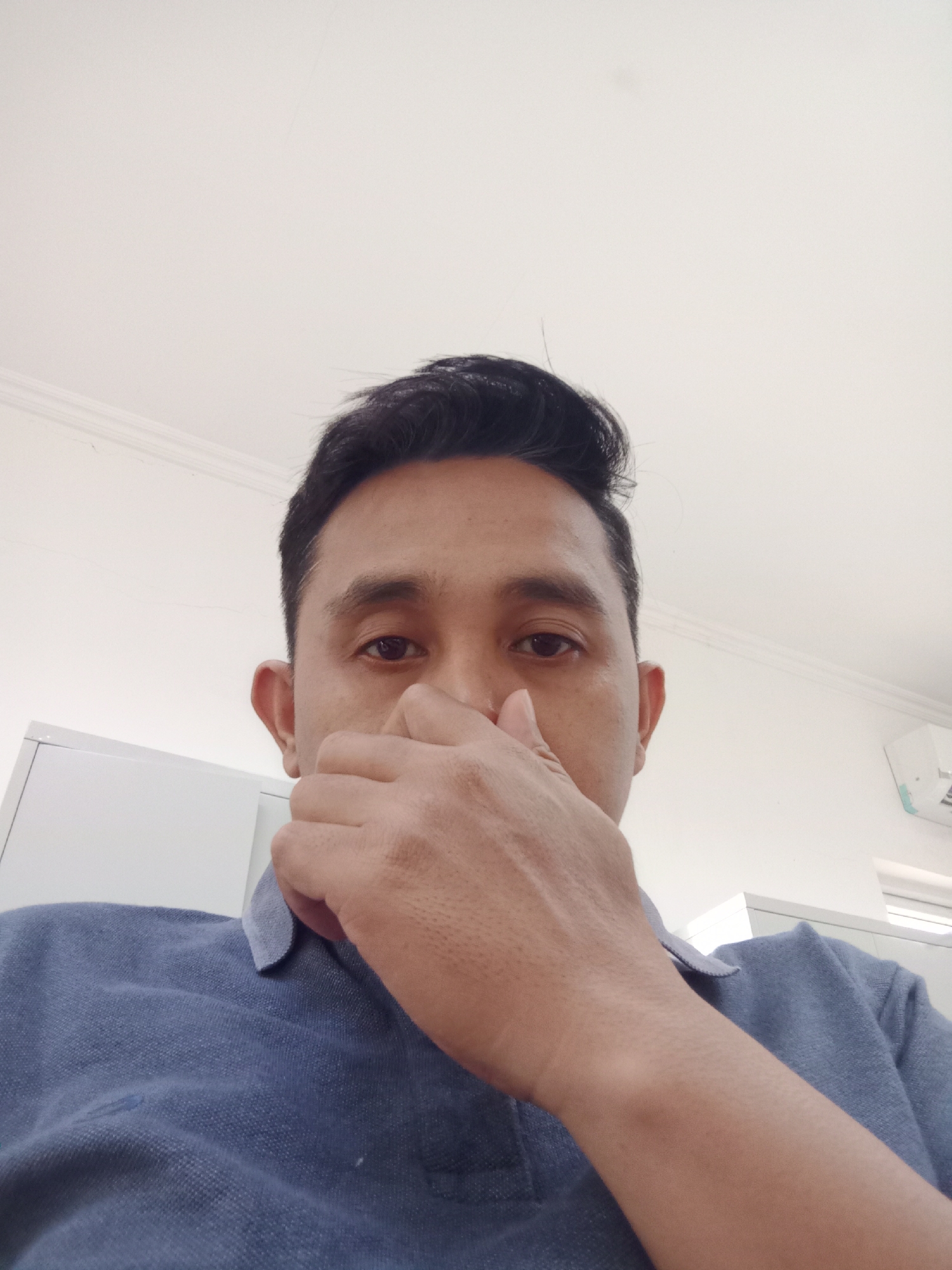 A low-angle, indoor, selfie-style, medium shot shows a man from the chest up. He is looking directly at the camera, and his right hand is covering his nose and mouth. The man has dark hair styled in a pompadour, brown eyes, and fair skin. He is wearing a blue polo shirt. The background is a plain white wall with a white ceiling at the top. The lighting is bright and diffused. The scene appears to be an office or similar indoor environment.