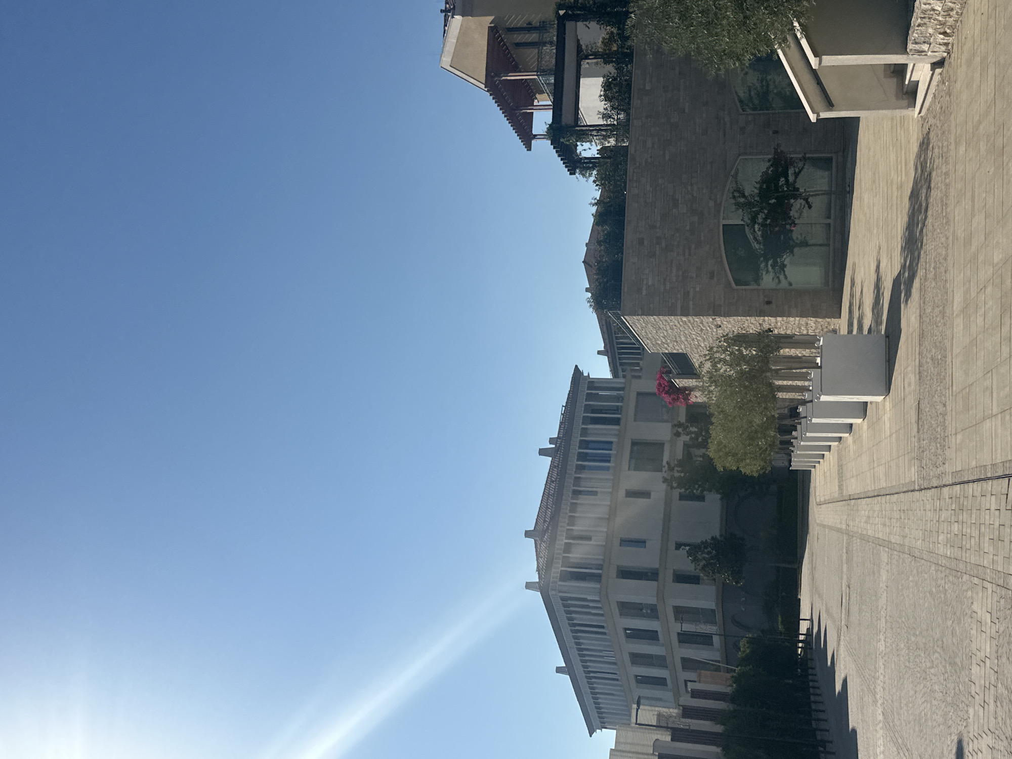 The image captures a sun-drenched, modern urban landscape in Kumbor, Montenegro. The scene is dominated by a clear blue sky with prominent sun rays stretching across the upper left portion of the frame. In the foreground, a paved walkway with a patterned brick design leads towards several contemporary buildings. On the right side of the walkway, a row of tall, white planters containing small trees is visible.
The buildings themselves are varied in architectural style. To the left, a large, multi-story building with a distinctive colonnade-like facade and a tiled roof stands prominently. Further to the right, a two-story building constructed with stone blocks features large arched windows, reflecting the surrounding trees and sky. Adjacent to this, another building with an upper-level balcony supported by wooden pergolas is visible. The architecture suggests a well-maintained, possibly commercial or residential complex.
Greenery is present in the form of trees lining the walkway and shrubs integrated into the building designs. The overall impression is one of a bright, clean, and modern environment in a Mediterranean setting, characteristic of a developed area within Kumbor, Montenegro. No people are present in the image.