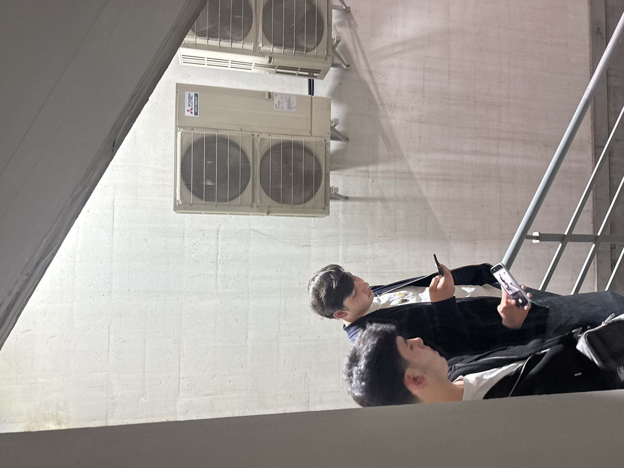 The image, taken from a low angle and oriented vertically, captures a scene likely within a modern building, parking structure, or industrial area in Yerevan, Armenia. The background features a light-colored concrete wall with a coarse texture, marked by subtle horizontal and vertical lines.
**Subjects:** Two men are present, viewed from a slightly elevated perspective. The man in the foreground, closer to the bottom of the frame, has dark, curly hair and a short beard, dressed in a white t-shirt and a dark jacket. His head and shoulders are visible. Behind him, and slightly higher in the frame, is another man with dark hair, wearing a white t-shirt with a graphic design and a dark jacket.
**Objects:** Mounted prominently on the concrete wall are several large air conditioning condenser units. Two main units, each with two circular grilles covering fans, are stacked. A smaller, rectangular unit with a control panel is positioned between them. To the far right, a light-colored metal railing, possibly part of a staircase or barrier, runs diagonally upwards, casting long shadows on the wall. Both men are holding smartphones. The man in the background also appears to be holding a small dark object, possibly a vape pen.
**Activity:** Both men are engrossed in their smartphones, looking down at their screens. They appear to be resting or taking a break in this setting.
**Notable details:** The lighting is even, suggesting an indoor or covered outdoor space, or an overcast day, producing soft shadows. The visible text on the air conditioning units clearly states "MITSUBISHI ELECTRIC" and "CITY MULTI." The overall ambiance is neutral, with the men focused on their personal devices rather than interacting with each other or their surroundings.