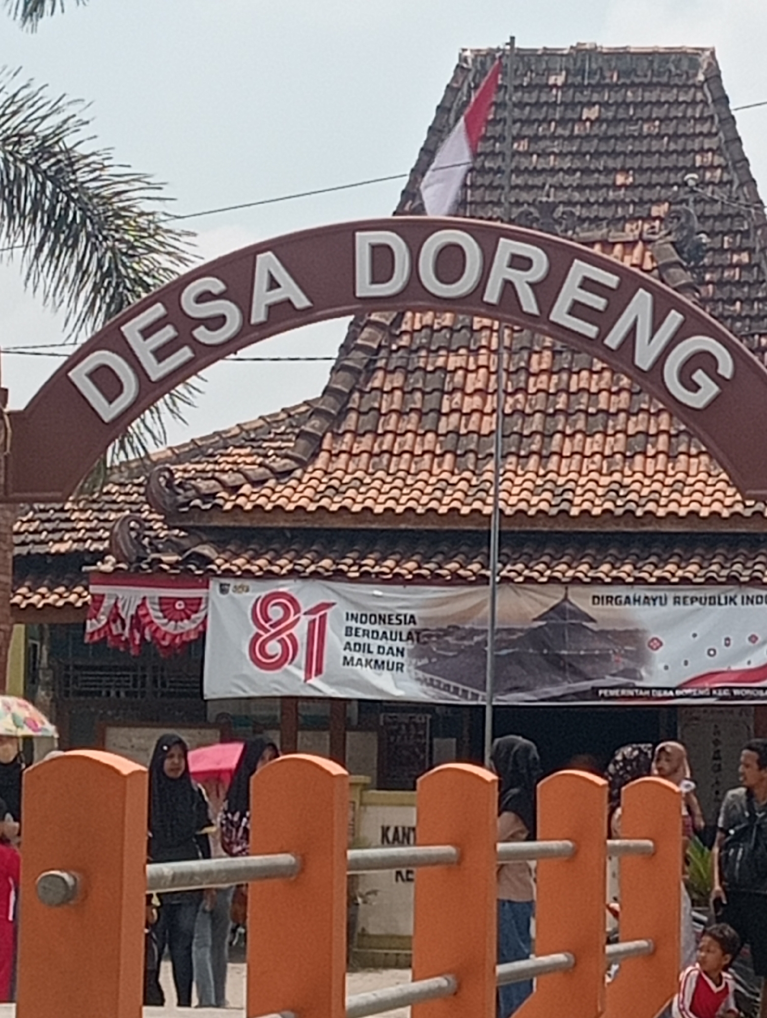 The image shows an outdoor scene in Doreng, Indonesia, with a prominent archway bearing the text "DESA DORENG." A red and white Indonesian flag flies from the roof of a building behind the archway, which is topped with traditional tiled roofing. Below the archway, a banner reads "81 INDONESIA BERDAULAT ADIL DAN MAKMUR" and "DIRGAHAYU REPUBLIK INDONESIA," with smaller text indicating "PEMERINTAH DESA DORENG KEC. WONOSARI."
Several people are visible in the foreground. Two women in hijabs stand behind an orange barrier. One woman wears a black hijab and dark clothing, while another wears a patterned hijab and a light-colored top. A third woman, also in a hijab, is further to the right, carrying a baby. A young boy in a red and white shirt is in the lower right corner of the frame. Other individuals are partially visible, and a child's umbrella is to the far left. The overall impression is of a community gathering or event in a public space within the village of Doreng.