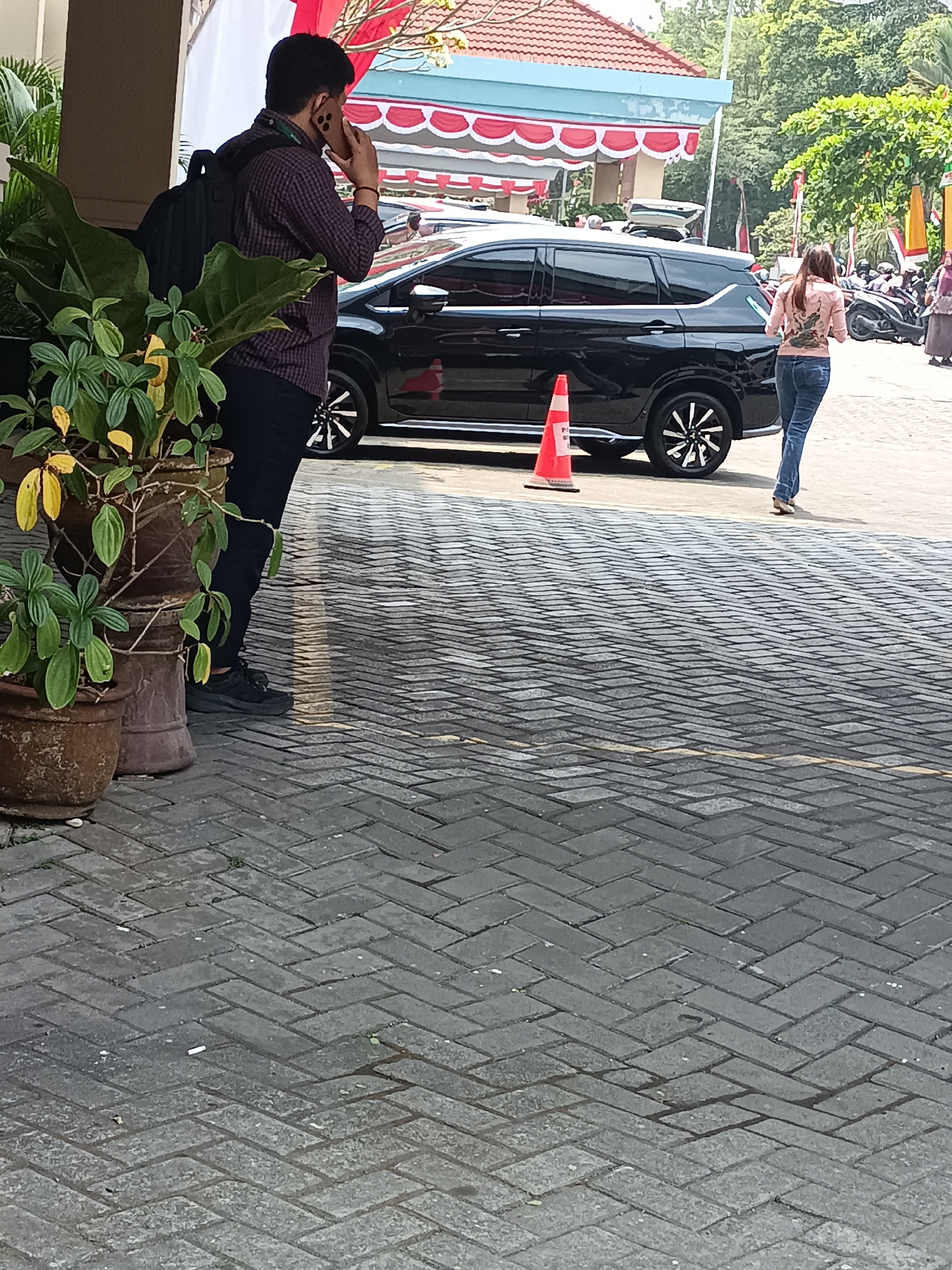 A man with dark hair and a plaid shirt is standing outdoors, talking on a cell phone. He is wearing a backpack and is positioned to the left of a black vehicle. In the foreground, there are several potted plants, and the ground is paved with interlocking gray bricks. A bright orange traffic cone is visible near the vehicle's rear wheel. To the right, a woman with long brown hair, wearing a light-colored jacket and jeans, is walking away from the camera. The background shows trees and buildings, suggesting an urban or suburban setting. The image appears to be taken in Sleman, Indonesia, likely in a public area such as a parking lot or walkway.