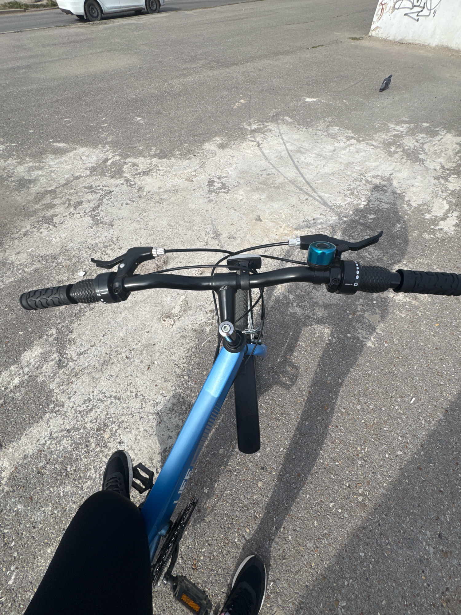 The image shows a first-person perspective of someone riding a blue bicycle on an asphalt road in Ufa, Russia. The road is somewhat worn and has patches of lighter colored concrete. To the left, a white car is partially visible parked on the side of the road. In the background, there is a white wall with graffiti. A pigeon is visible on the ground to the right. The handlebars of the bicycle are black, with black grips, brake levers, and a blue bell. The rider is wearing black pants and black athletic shoes. The shadow of the bicycle and rider stretches across the road, indicating it is a sunny day.