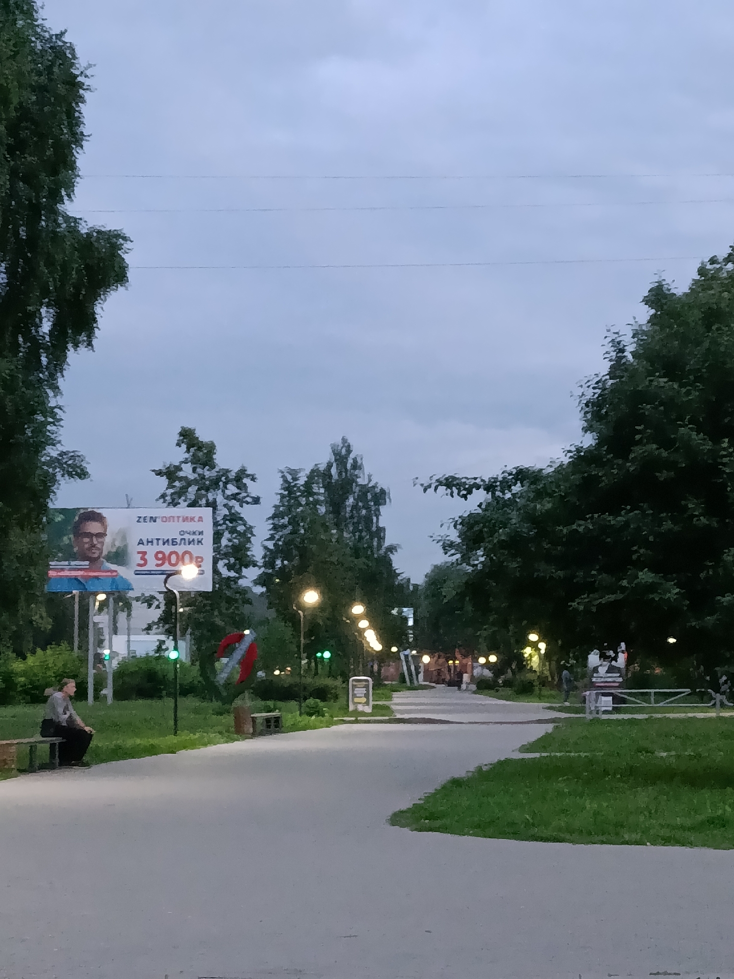 This is a photograph taken on a cloudy day in Chaikovsky, Russia, likely in the evening given the artificial lighting visible. The scene depicts a park or public walkway lined with trees. In the foreground, a paved path curves through green grass. A person is seated on a bench on the left side of the path. Further down the path, streetlights illuminate the walkway. A large billboard is visible on the left side of the image, advertising "ZEN OPTIKA" for "ANTI-BLIK" glasses, priced at 3,900 rubles. Another billboard or sign is visible on the right side of the path. The overall atmosphere is calm and overcast.