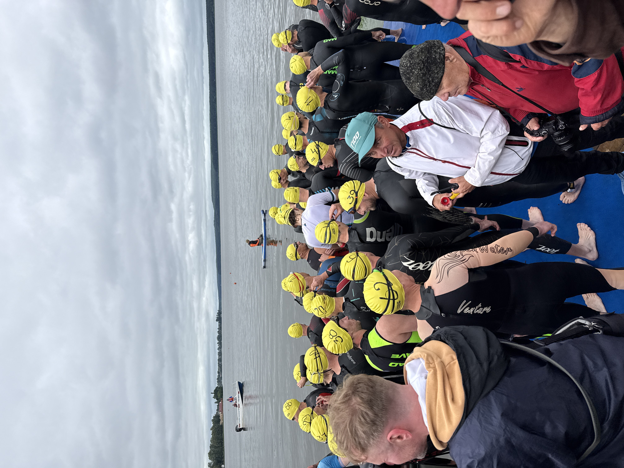 This is a high-angle, eye-level, full-shot photograph of a group of swimmers gathered on a blue platform near a body of water. The swimmers are mostly wearing black wetsuits and yellow swim caps. Many of the caps have numbers on them, such as "66", "38", and "11". Some swimmers have tattoos on their arms. In the background, there is a calm lake with a distant shoreline lined with trees. A small white boat with people in it is visible on the water, as well as a person on a paddleboard. The sky is overcast with gray clouds. Two men in the foreground are engaged in conversation. One man is wearing a white jacket with red trim and a blue baseball cap, and he is holding a small yellow object. The other man is wearing a red jacket and a dark knitted hat, and he is holding a camera. The scene appears to be part of an outdoor sporting event, likely a triathlon or swimming race. It is plausible this is in Votkinsk, Russia, given the context of the provided information, though specific landmarks to confirm the location are not visible.