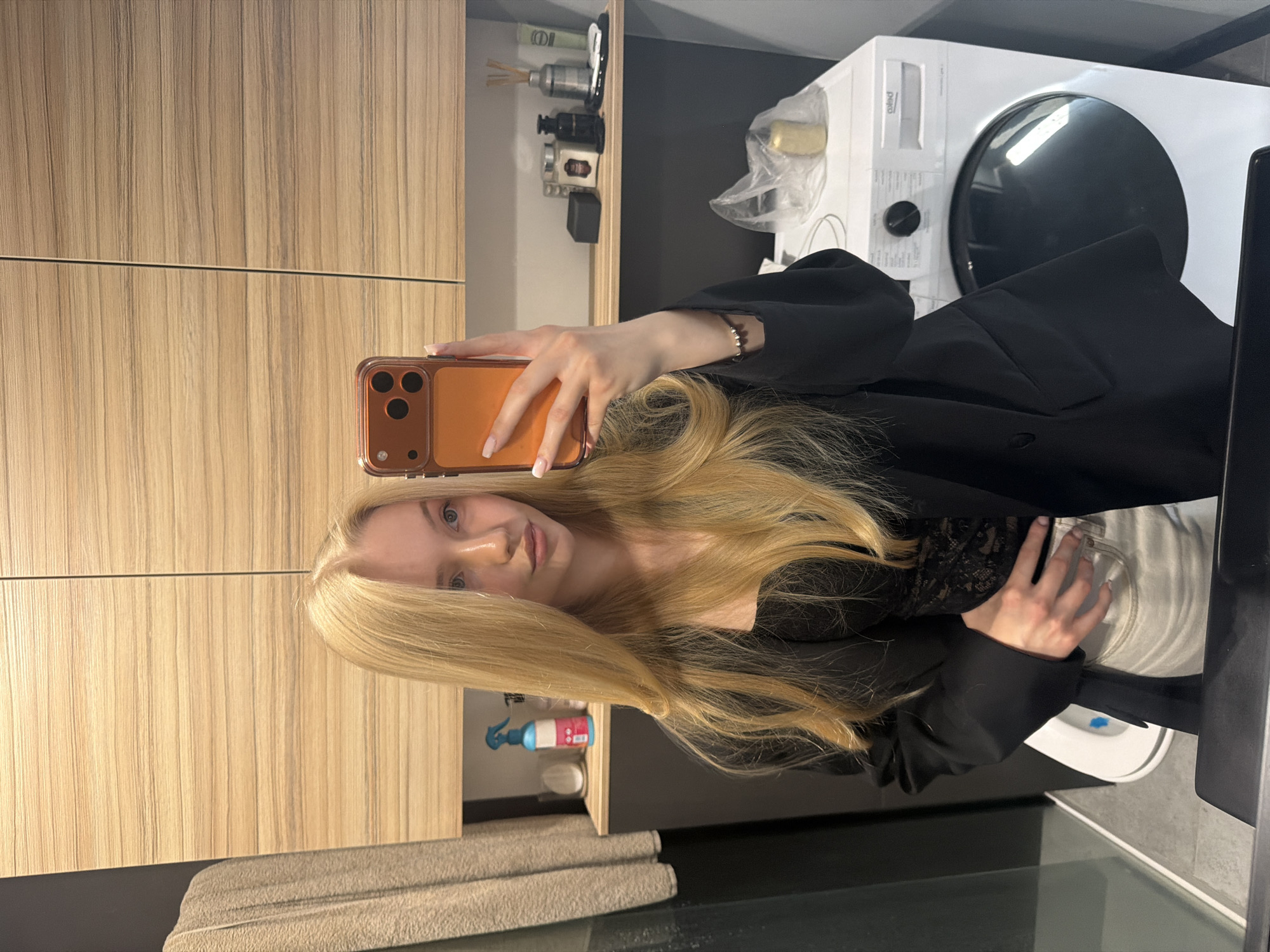 A young woman with long blonde hair is taking a selfie in what appears to be a bathroom. She is holding an orange phone in her right hand. She is wearing a black blazer over a black top with lace details. The bathroom features light-colored wooden cabinets on the left side of the frame and dark gray walls. On a shelf above a gray towel, there are cleaning supplies and a white container with a blue spray nozzle. To the right, a white washing machine is visible. A partial view of what appears to be a toilet and other bathroom fixtures can be seen in the lower right corner of the image. The location is Unknown, Unknown.