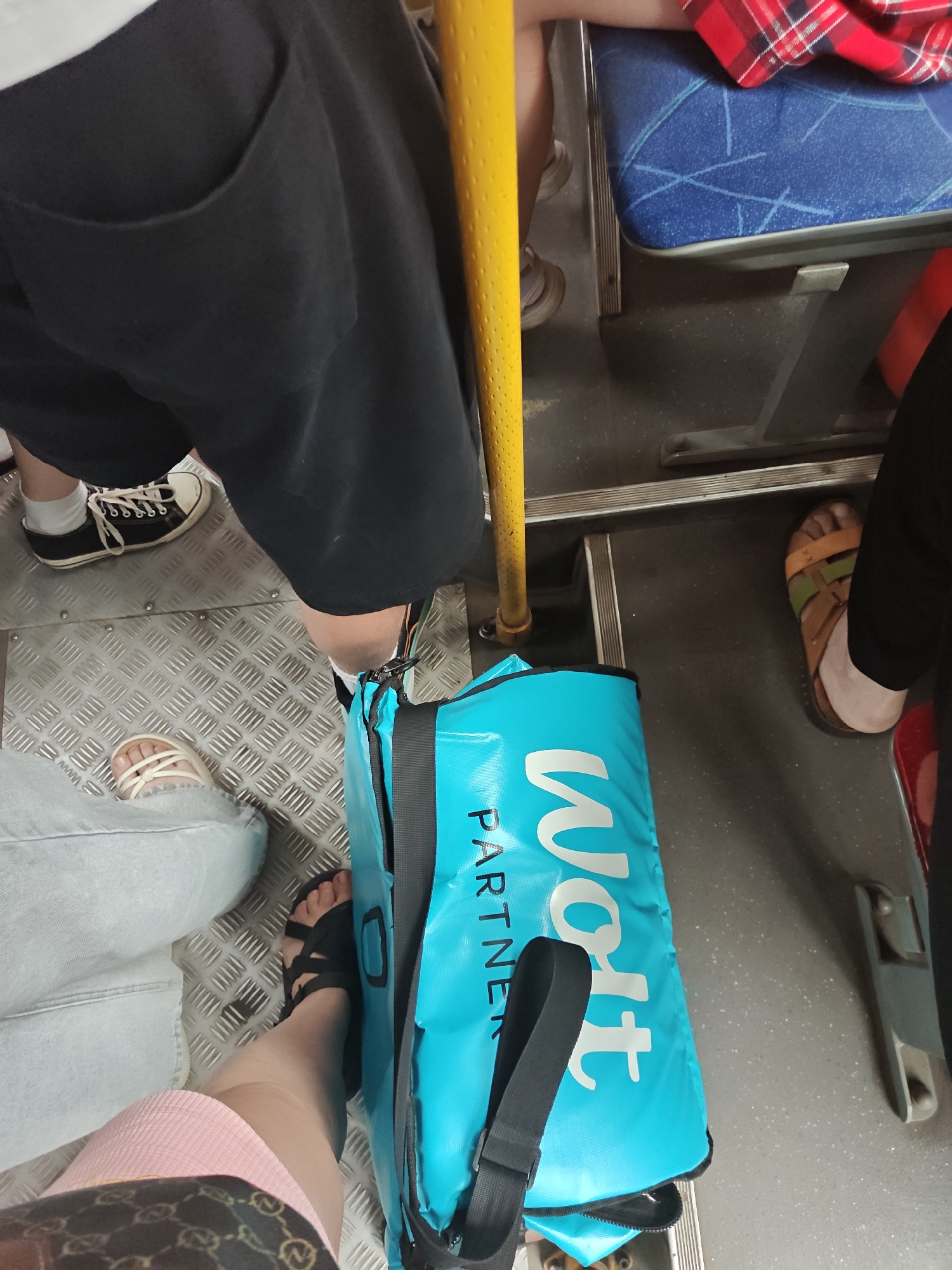 The image depicts the interior of a public transport vehicle, likely a bus, with multiple individuals' legs and feet visible. The floor is made of textured metal. A bright yellow pole is a prominent vertical element on the left side of the frame. A large, turquoise bag with white lettering that reads "Wolt Partner" is positioned on the floor. Various people are wearing casual footwear, including sneakers, sandals, and flip-flops. One person is wearing black shorts, another is wearing light-colored jeans, and a third is wearing a pink skirt. The seats appear to be blue with a patterned design. In the background, a red and black plaid fabric is visible on a seat. The overall scene suggests a crowded mode of transportation. There is no visual information to indicate the location is specifically Petropavl, Kazakhstan.