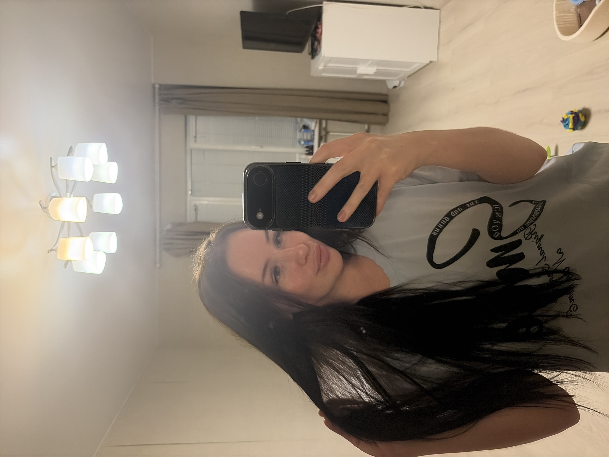 A mirror selfie captures a young woman in an indoor setting, likely an apartment in Saint Petersburg, Russia. The image is rotated 90 degrees clockwise. The woman, with long dark hair flowing over her right shoulder and down her chest, has a light complexion and a soft smile, showing faint dimples. She is wearing a light-colored t-shirt with dark, stylized text partially visible on the front; decipherable parts include "around the world", "LICE", and "SO". Her left hand holds a black smartphone with a textured case, angled towards the mirror to take the photograph.
The background, reflected in the mirror, reveals a modern room. To the left, a multi-bulb chandelier or wall light fixture emits a warm glow from several white, cylindrical shades. Further in the background, a flat-screen television is mounted on a wall, and below it, a white cabinet or appliance, possibly a mini-fridge. A window with closed blinds is also visible. The floor is made of light-colored wood, and a small, colorful, spherical toy can be seen on it. The walls are a neutral light color. The overall lighting suggests it is evening or night, relying on artificial light sources. The atmosphere appears casual and comfortable.
