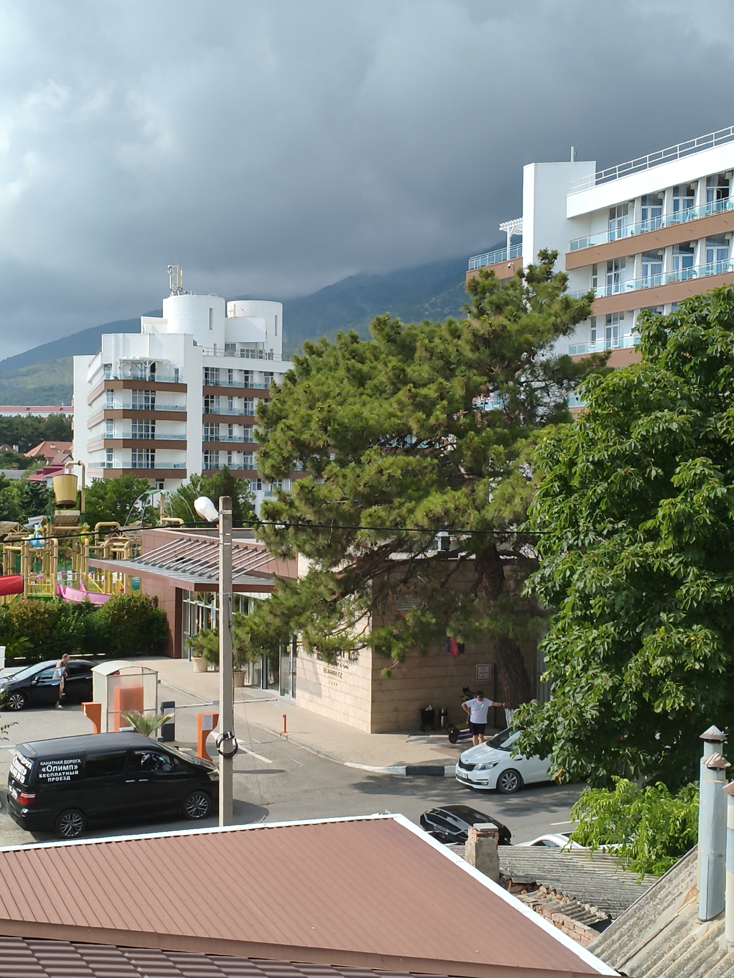 The image depicts a view of the "Olimp" hotel in Gelendzhik, Russia, on a cloudy day. In the foreground, a brown tiled roof is visible. Behind it, a black minivan with text in Russian is parked. To the left, a children's playground with slides and water features is partially visible. Further back, a tall white building, the "Olimp" hotel, dominates the scene, with multiple floors featuring balconies and windows. A large evergreen tree stands in front of the hotel, obscuring part of its facade. In the middle ground, a man in a white t-shirt and dark shorts stands near a white car. Mountains are visible in the background under a cloudy sky.