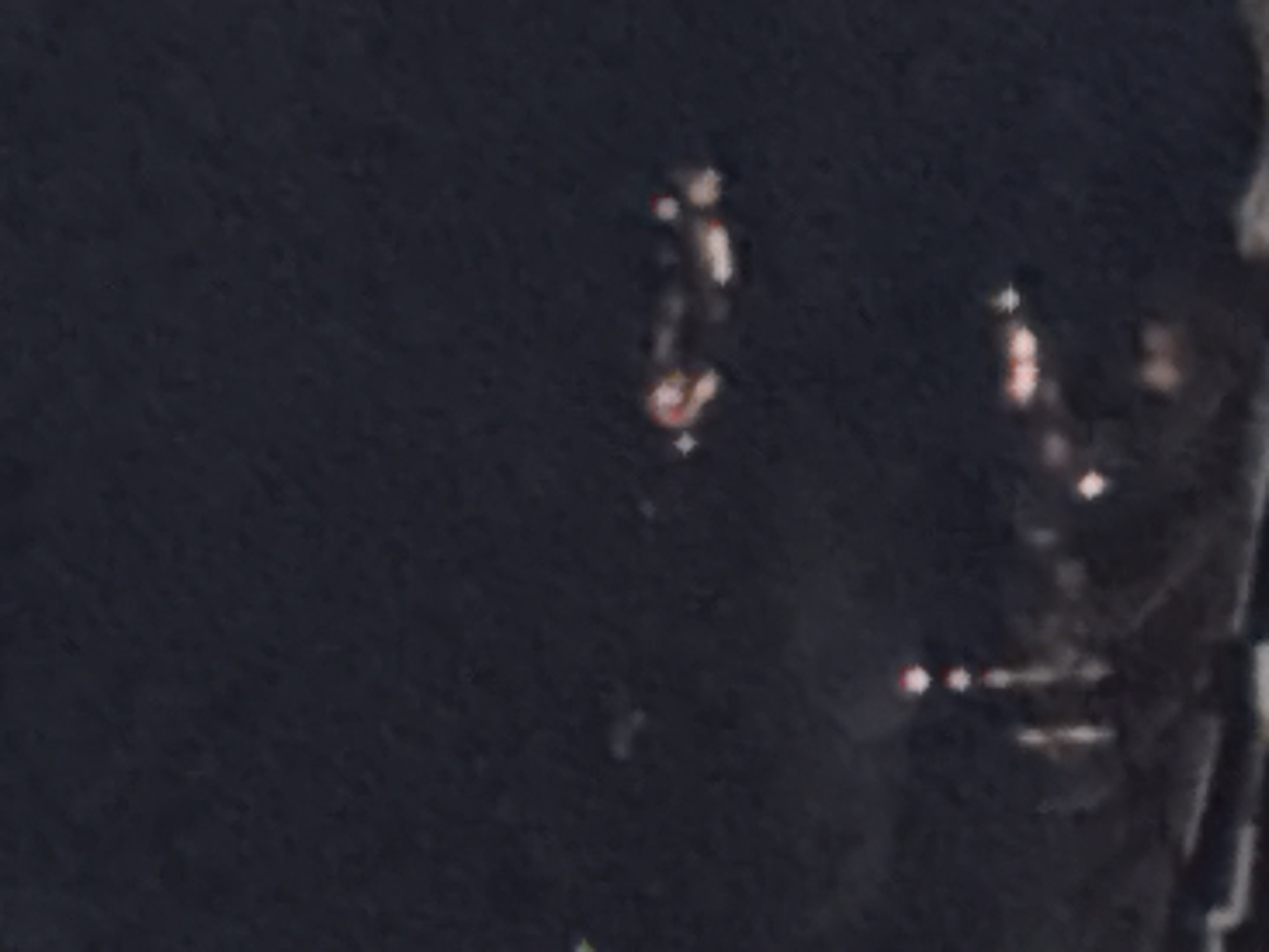 The image is a nighttime aerial or elevated view of a city, possibly Boreti, Montenegro, characterized by numerous blurred lights. In the upper right quadrant, a distinct object with multiple lights, appearing to be a vehicle or aircraft, is visible. Below this, in the lower portion of the frame, are more lights, some arranged in vertical patterns that might indicate buildings or structures. The overall scene is dark, with the lights providing the primary illumination and detail. The resolution is low, and the image is grainy, making precise identification of smaller elements difficult.