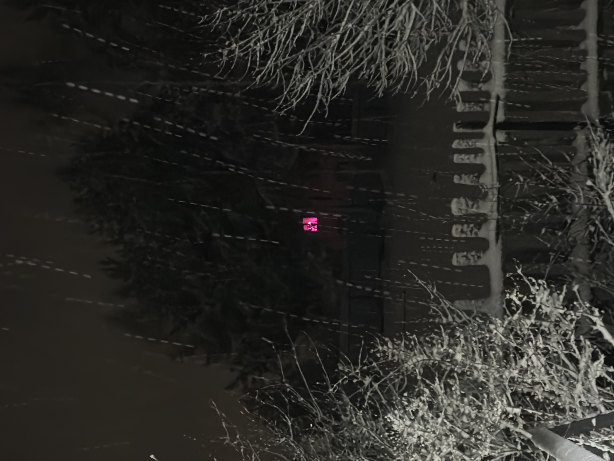 A monochrome, nighttime image shows a snow-covered scene in Markovo, Russia. The view appears to be from a slightly elevated position, possibly a window. Snow is falling, creating streaks of white light across the dark background. Bare tree branches are visible in the foreground and at the top of the frame, also coated with snow. In the center, a dark, rectangular shape suggests a building, with snow clinging to its vertical edges. A small, bright pink or magenta object is visible on the building.