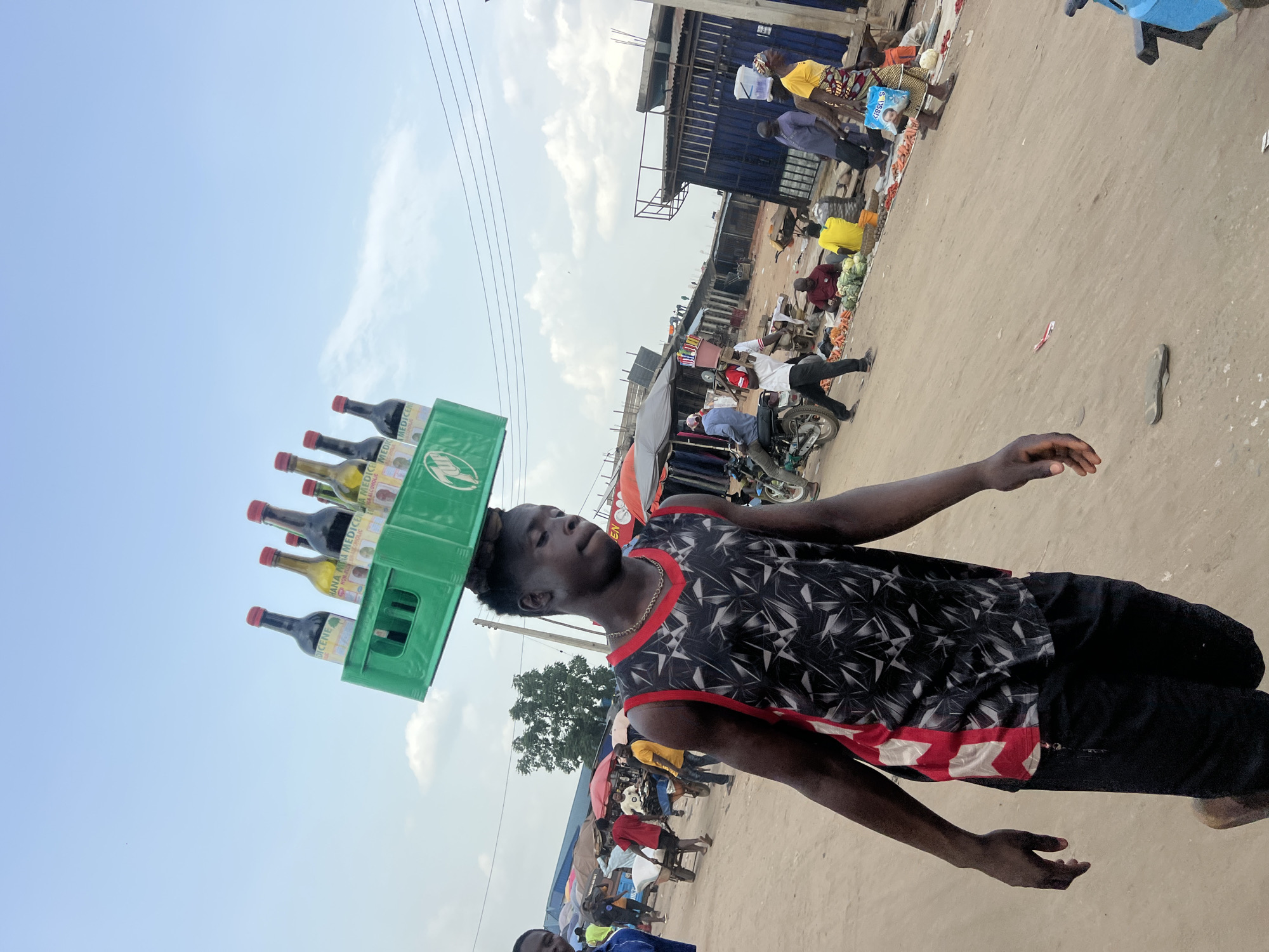 A young man balances a green crate of bottles on his head, navigating a bustling outdoor market scene in Oko Amakom, Nigeria. The crate contains bottles labeled "MEDICERE". He wears a black and white patterned tank top with red trim and dark pants. In the background, numerous people are engaged in market activities, with stalls and various goods visible. A motorcycle is parked nearby. Buildings and utility poles are visible in the distance under a partly cloudy sky. The ground is sandy and appears well-trafficked.