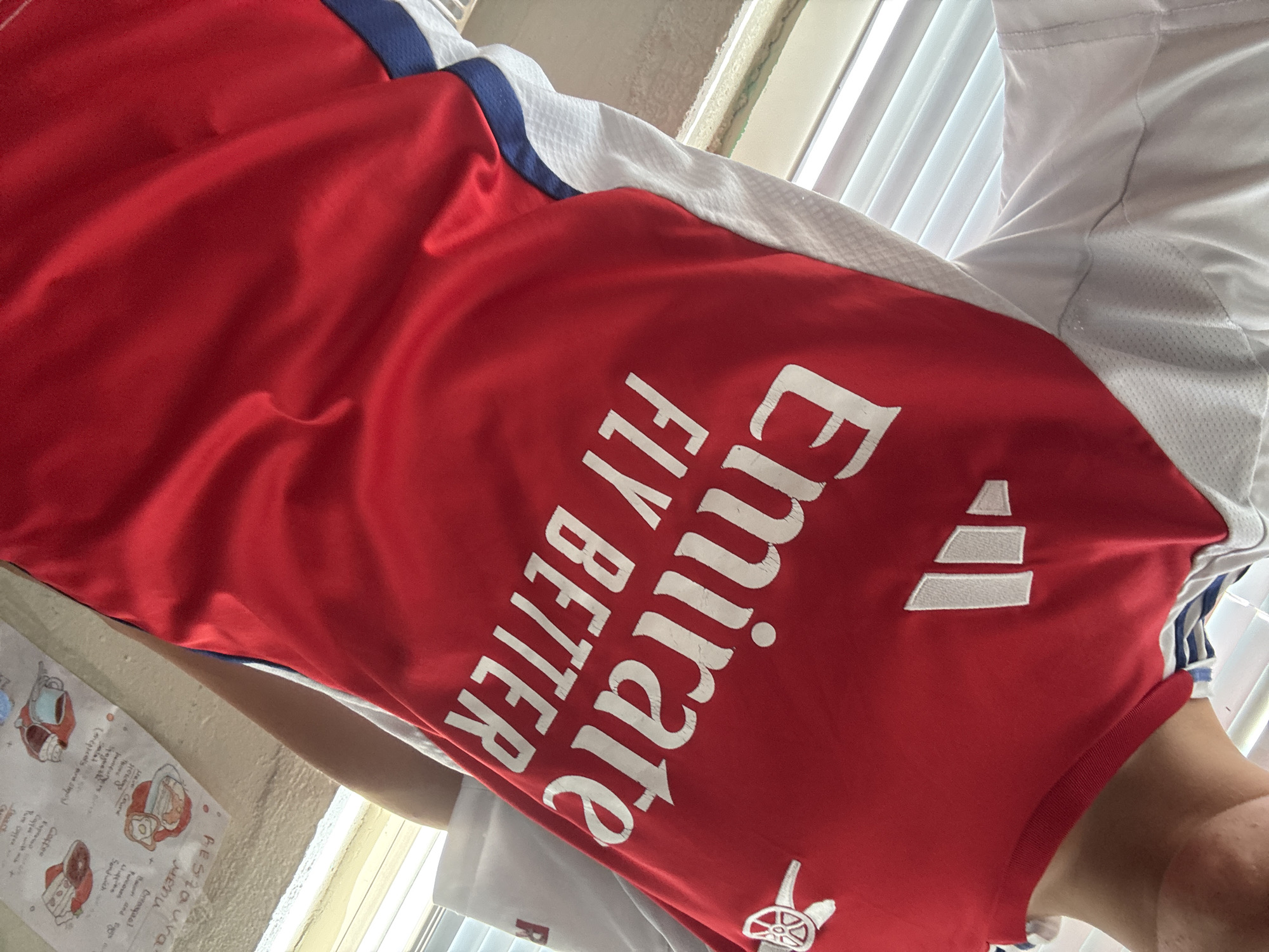 A person is wearing a red Arsenal football jersey. The jersey has white Adidas stripes on the shoulders and the Emirates Fly Better sponsor logo printed in white on the chest. A white Arsenal crest is visible on the upper left chest. The background shows a window with white blinds and a wall with a handwritten menu or poster. The scene is indoors, with natural light coming from the window.