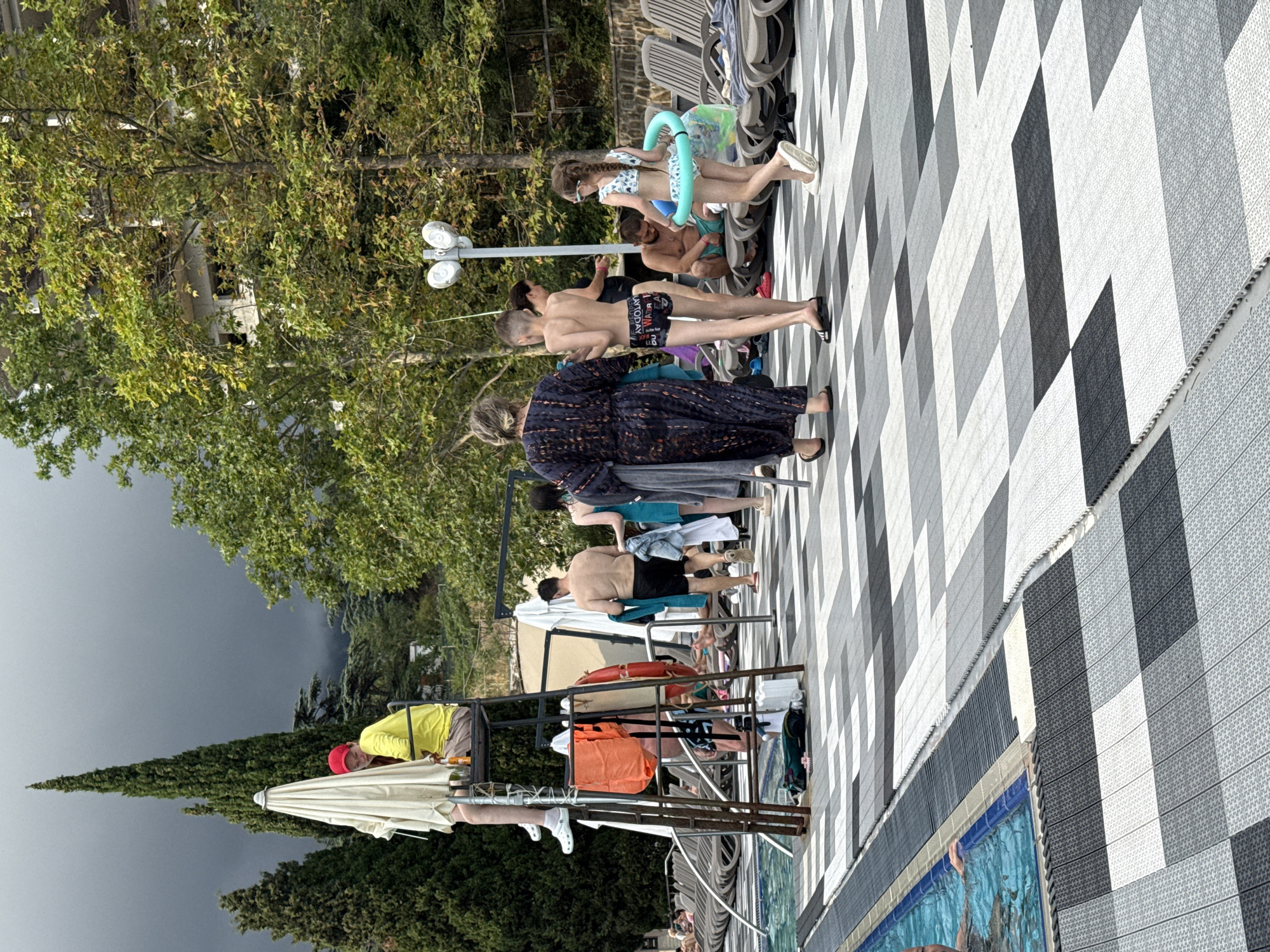The image depicts a scene at a swimming pool area, likely in Yalta, Ukraine, given the context. The weather appears overcast, with dark clouds visible in the background and tall, dark green cypress trees framing the scene. In the foreground, a black and white checkered tile pattern covers the ground. A lifeguard in a yellow shirt and red cap sits on a high lifeguard chair, with orange and red life preservers hanging from it.
Several people are present. To the left, partially in the pool, is an individual whose arm is visible. Further back, several individuals in swimwear are walking or standing around the pool area. Two children are prominent: a boy in dark swim trunks and a girl wearing a swimsuit and a blue inflatable ring. A woman in a dark patterned dress stands near the children. Another man in black swim trunks is seated on a lounge chair in the background. Numerous lounge chairs are arranged around the pool. A multi-story building, partially obscured by trees, is visible in the background.